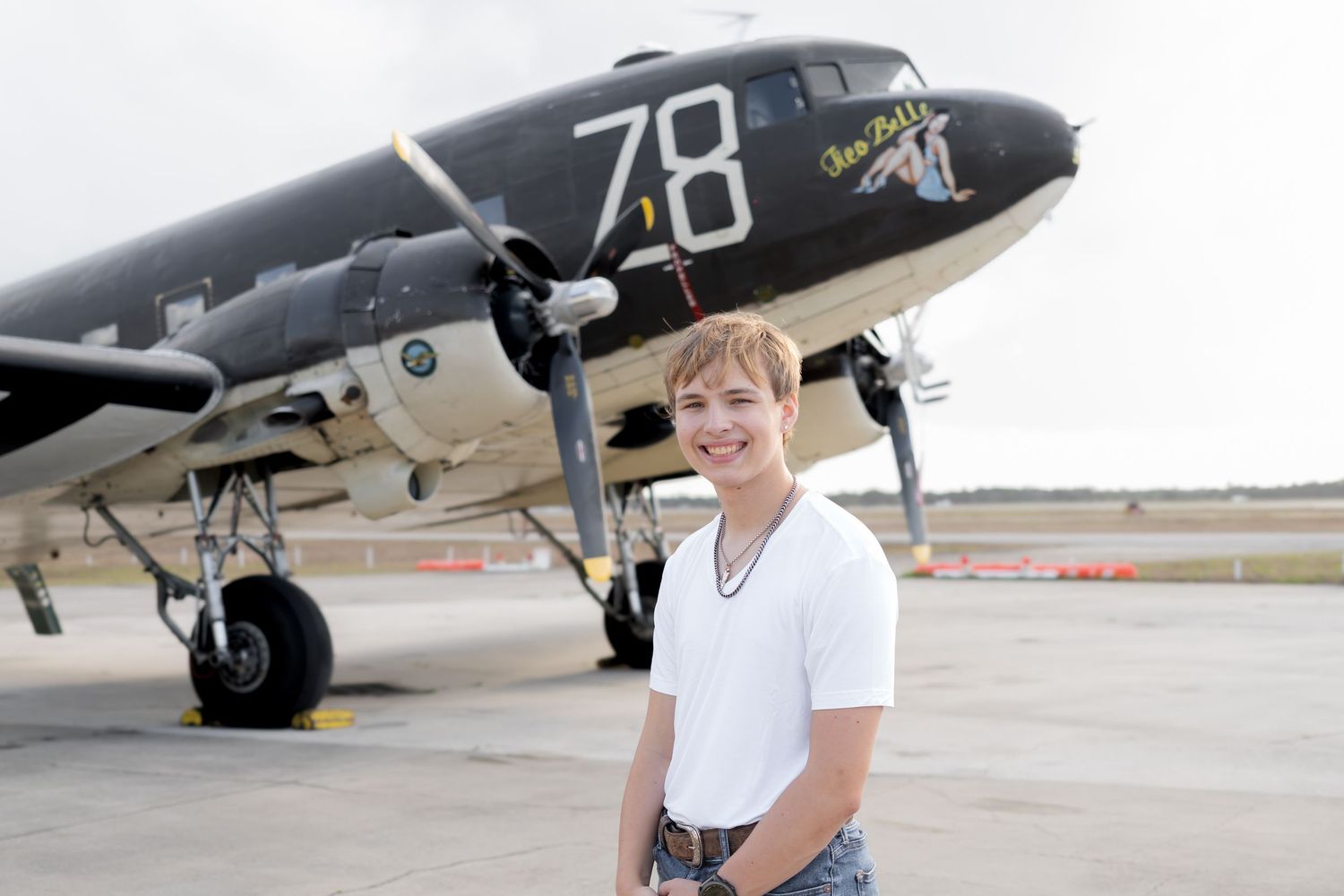 Young person smiling in front of a vintage WWII DC-3 aircraft numbered 78 on an airport tarmac.