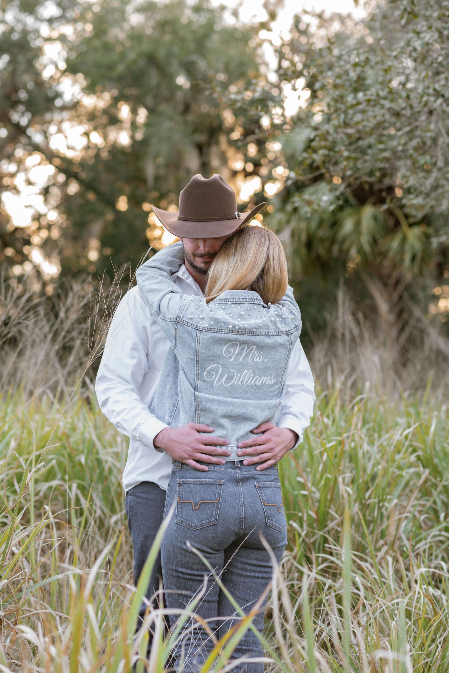 Couple embracing in tall grass field, woman wearing custom Mrs. Williams denim jacket, man in cowboy hat.