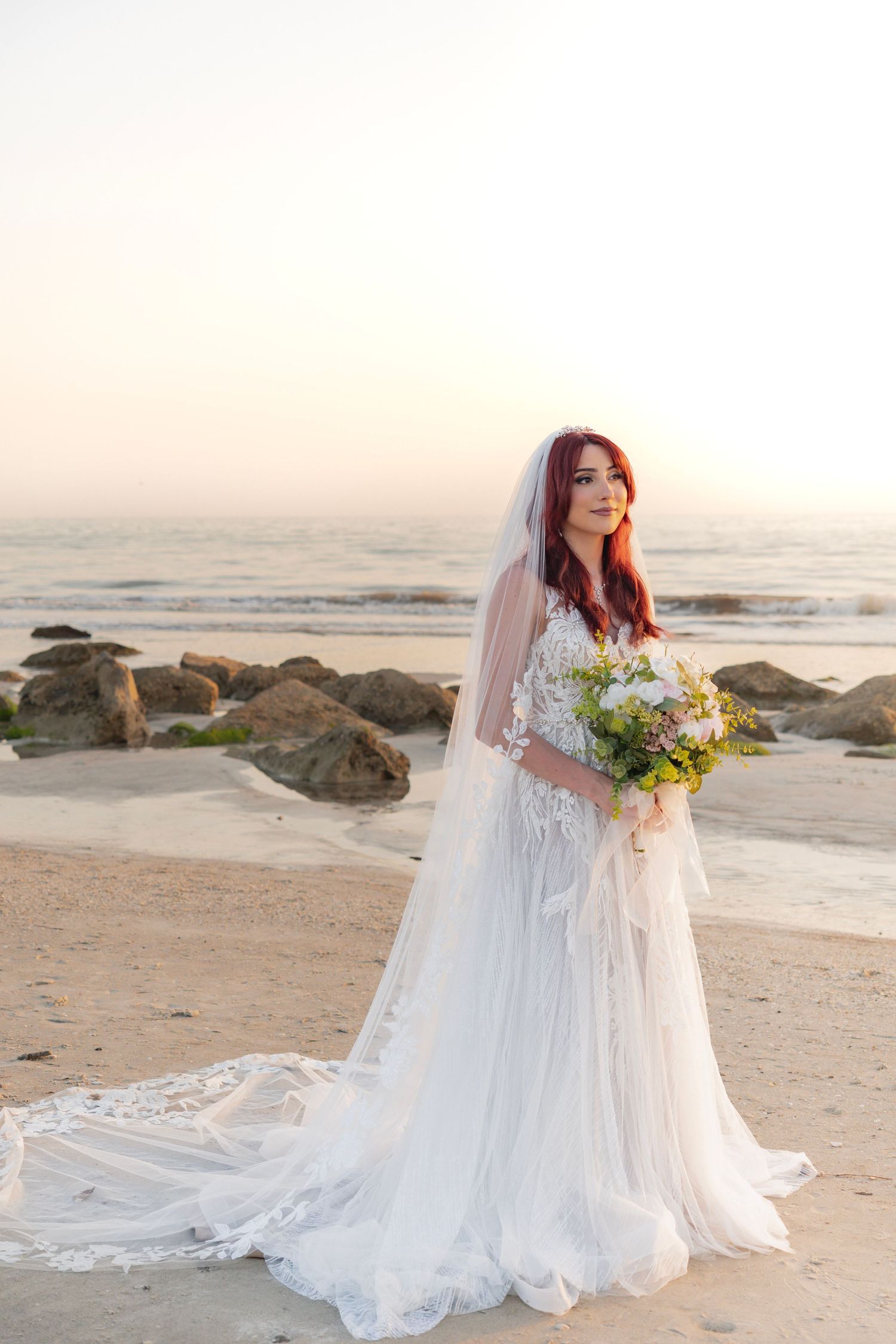 Bride in white lace gown with veil holds floral bouquet on sandy beach at sunset with rocks and ocean.