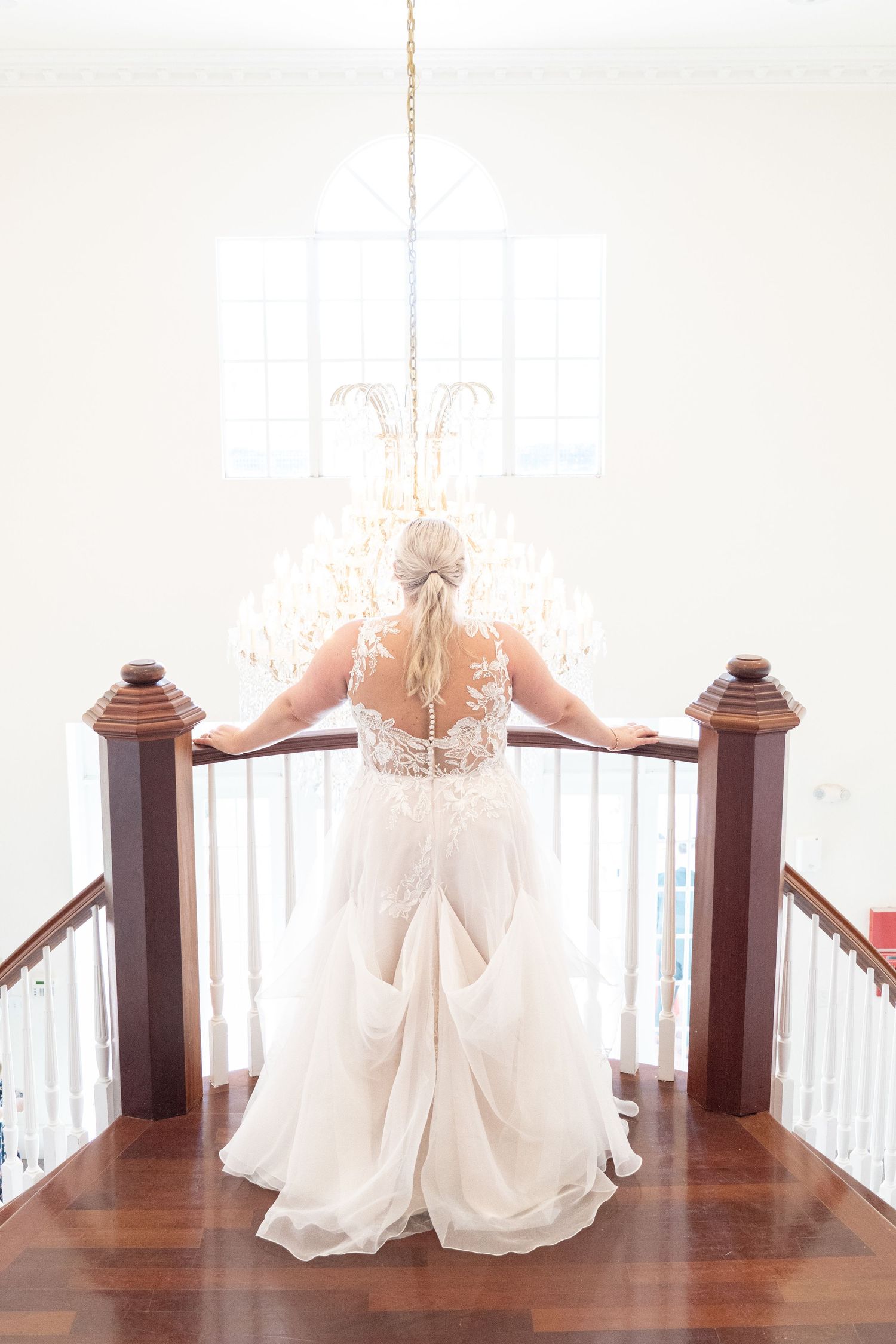 Bride in lace backless wedding gown stands at staircase railing beneath elegant crystal chandelier.