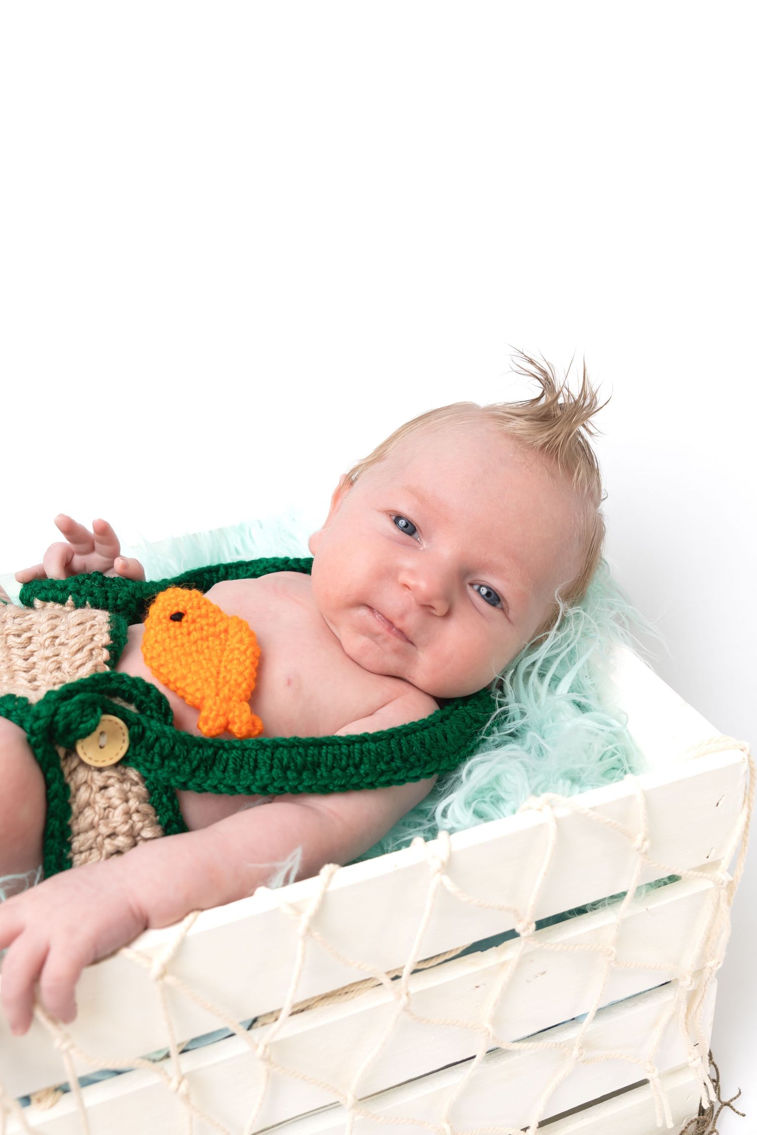 Cute baby in green knit overalls with orange chick toy lying in white wooden crate with teal shredded filler.
