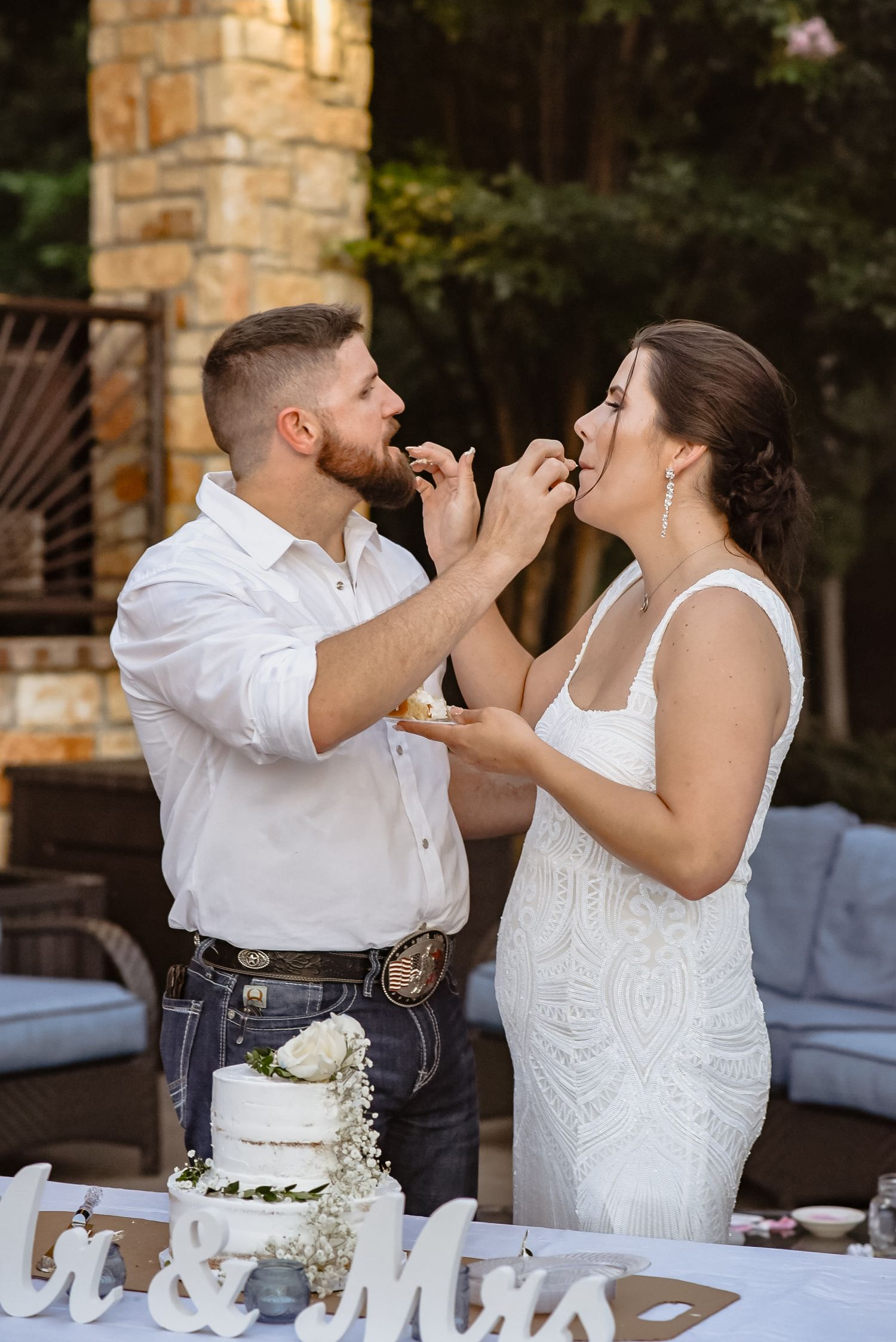 Bride and groom feeding each other wedding cake at outdoor reception table with white tiered cake and Mr. & Mrs. sign.