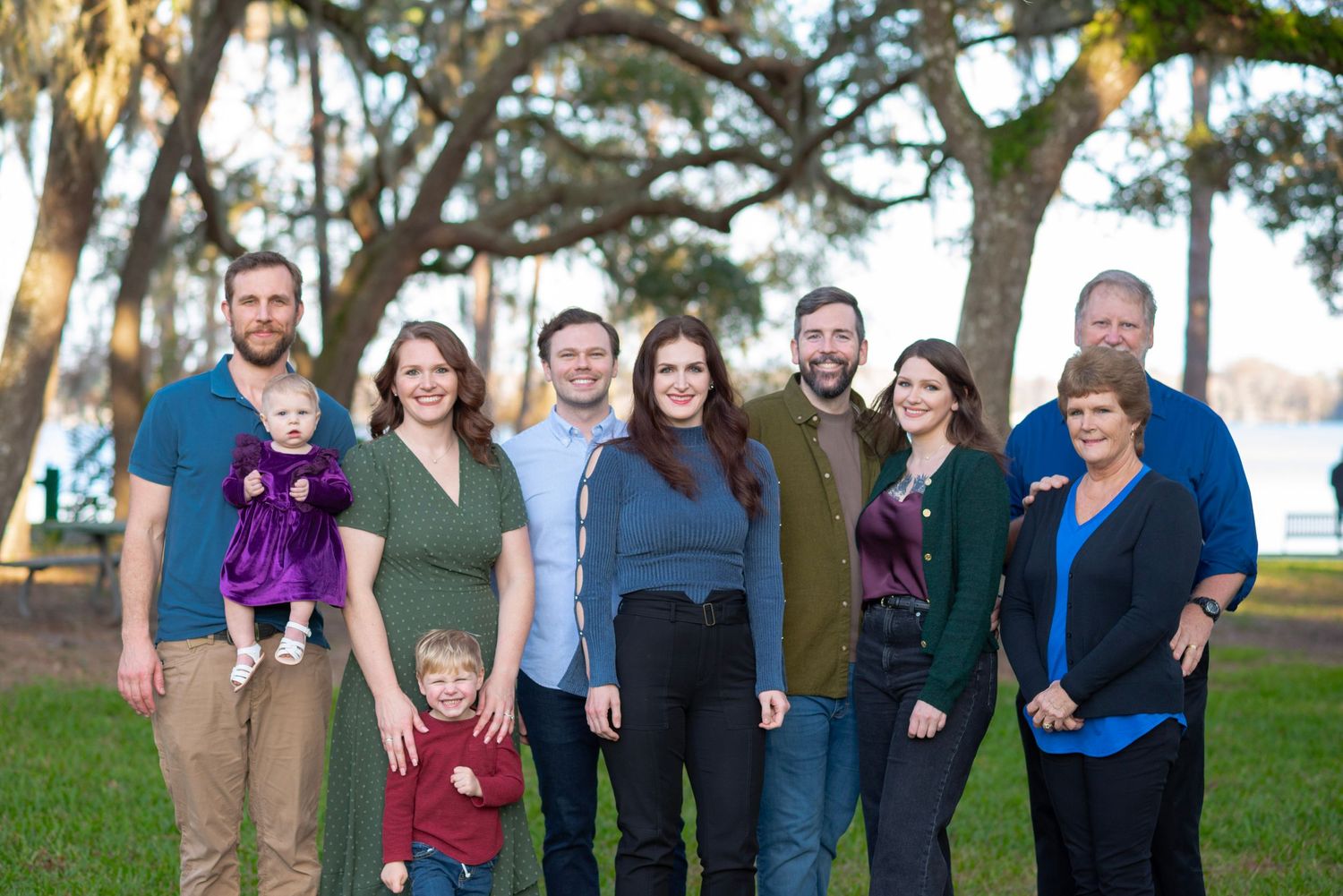 Large family group portrait outdoors in a park with moss-covered trees and a lake in the background.