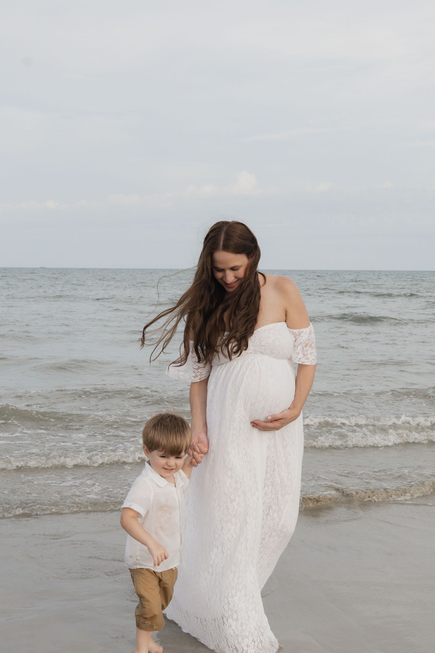 Pregnant woman in white lace off-shoulder dress walks on beach holding young boys hand near ocean waves.