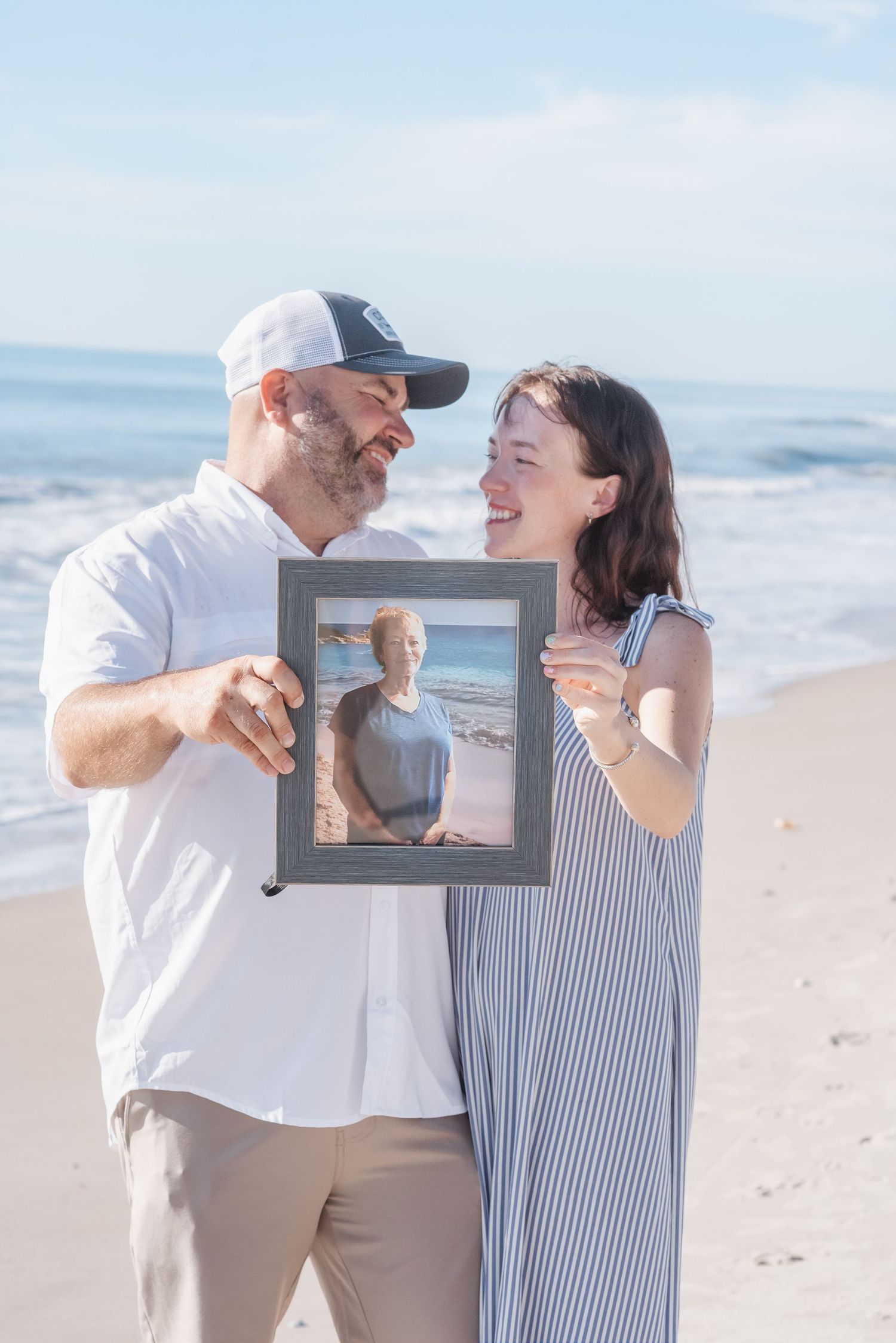 Couple smiling at each other on a beach while holding a framed photo, ocean waves in the background.