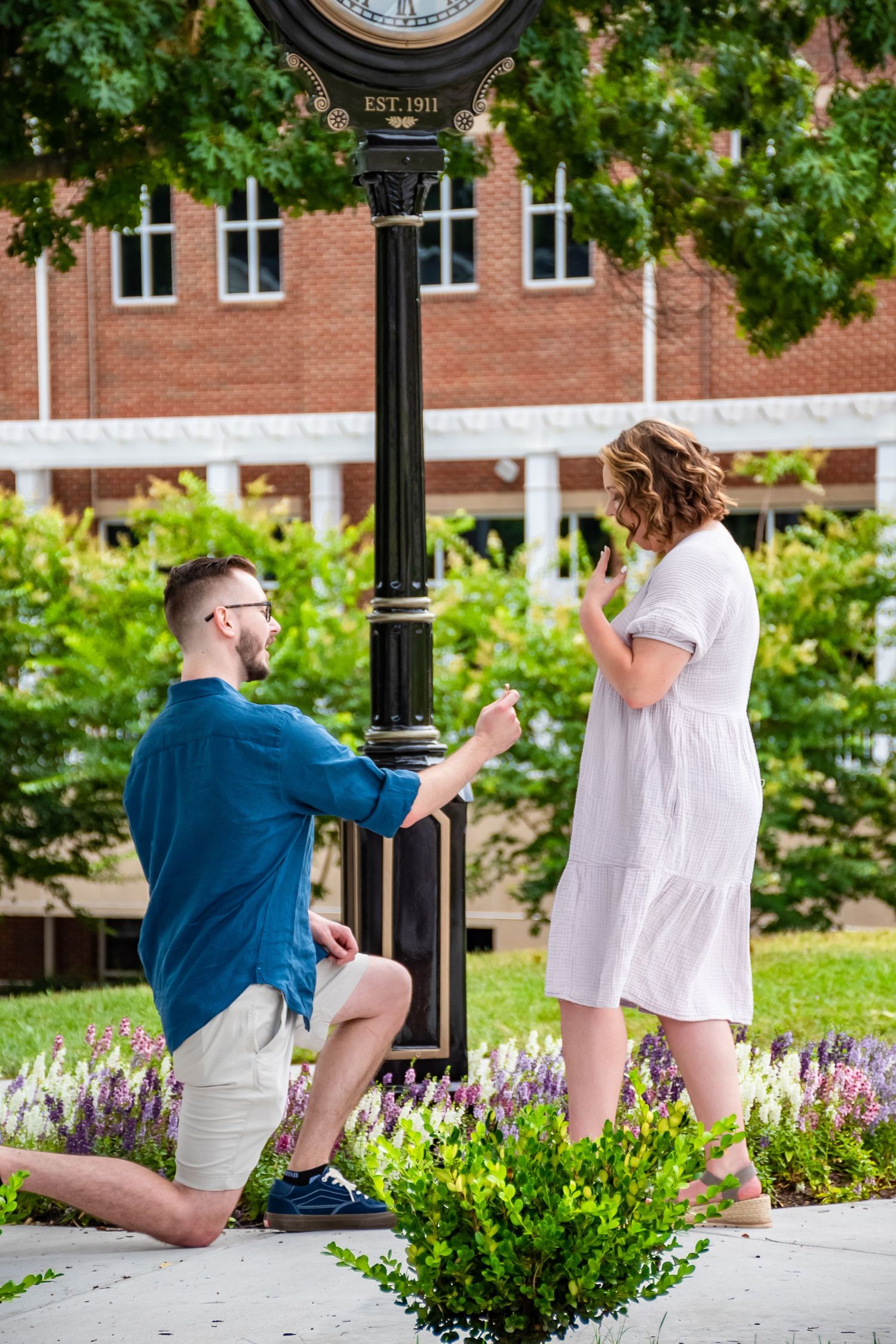 Man proposes on one knee to woman in white dress near ornate clock tower in lush garden setting.