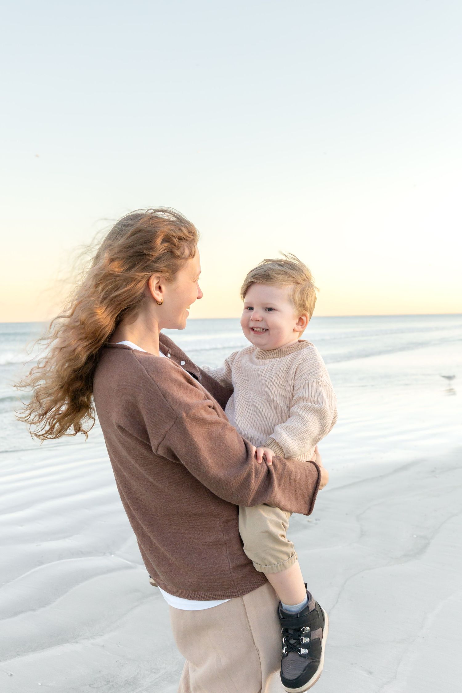 A young woman holds a smiling toddler on a white sand beach at sunset, both wearing neutral tones.