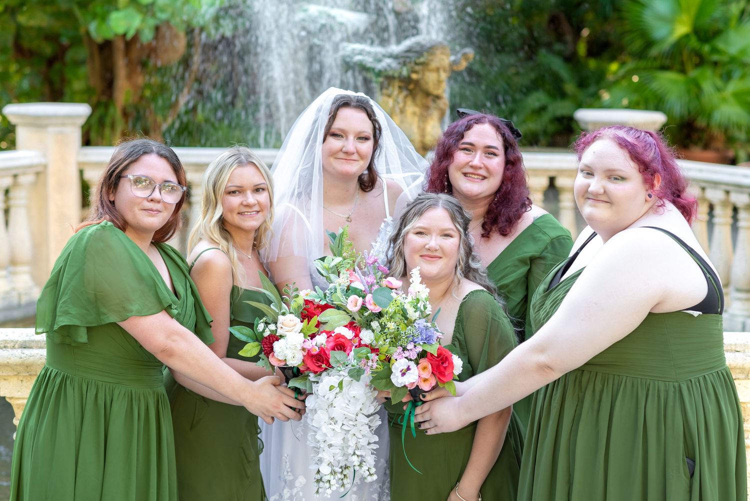 Bride in white gown and veil poses with five bridesmaids in green dresses holding bouquets near a fountain.