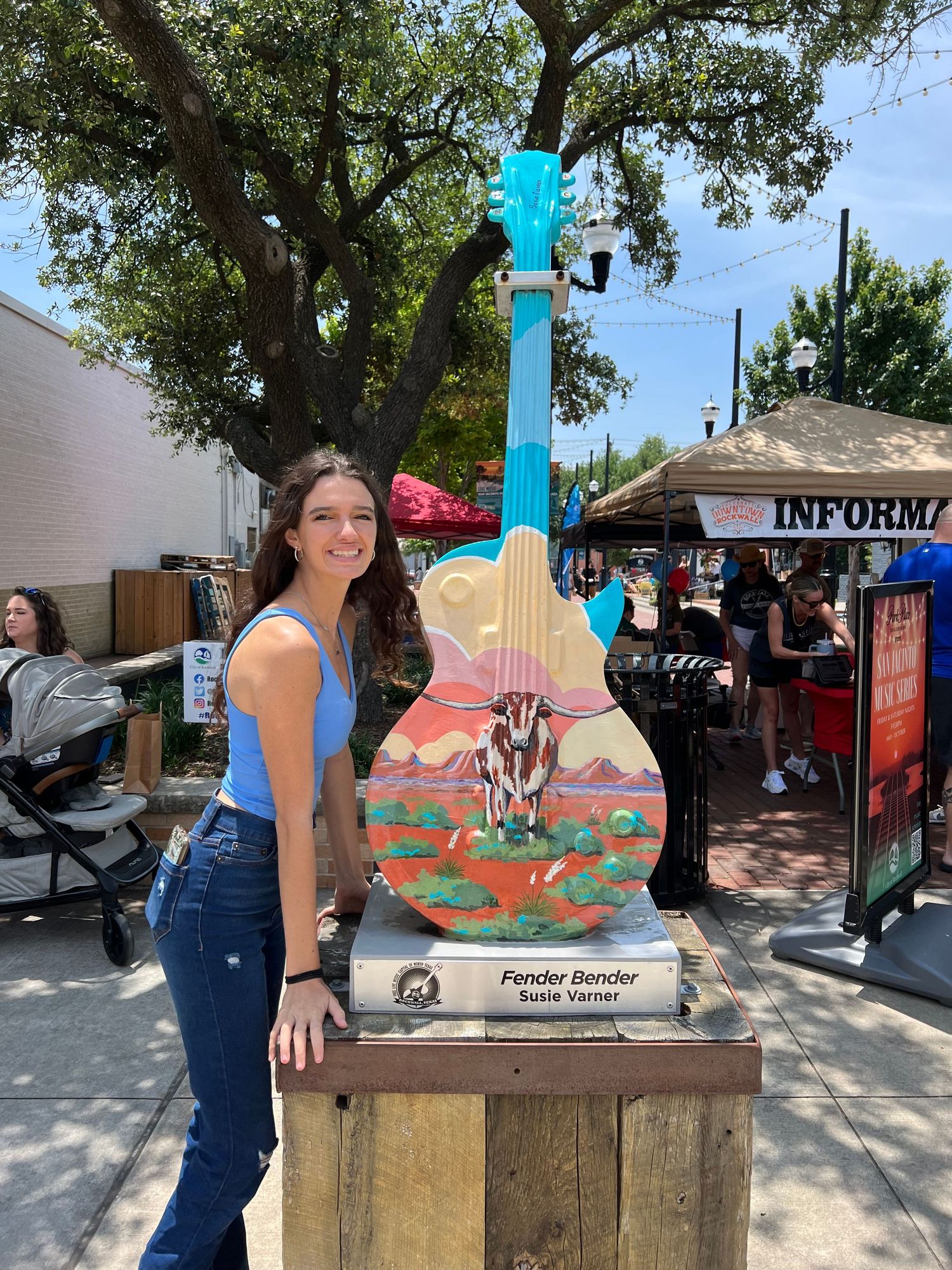 A woman smiles beside a colorful painted guitar sculpture at an outdoor market.