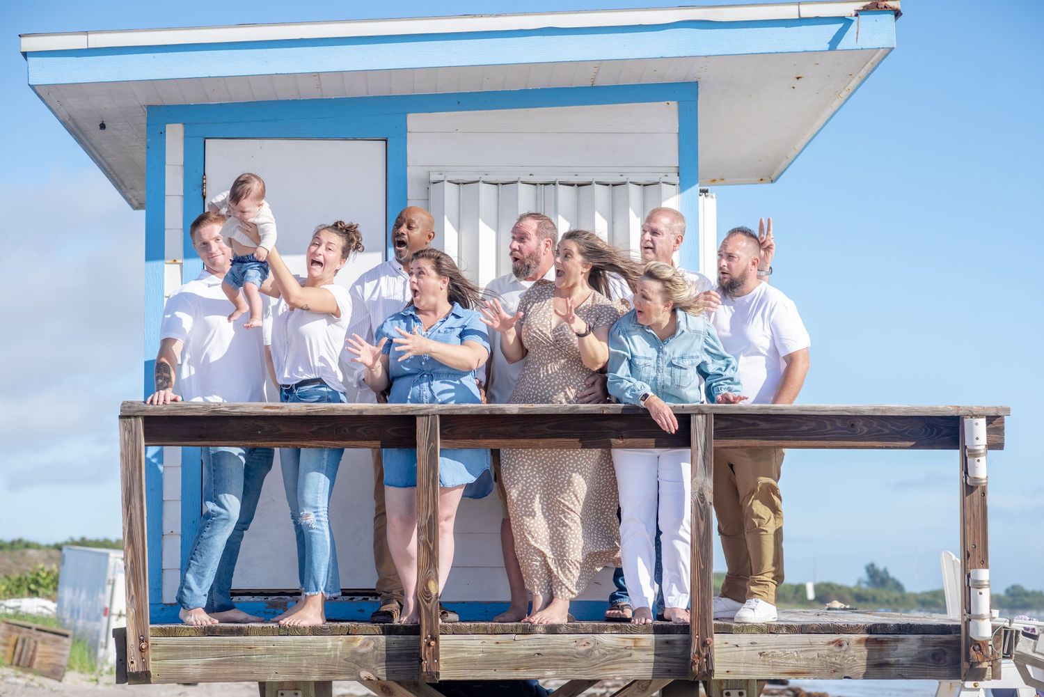 A joyful group of people laughing and posing on a wooden deck by a blue and white beach lifeguard hut.