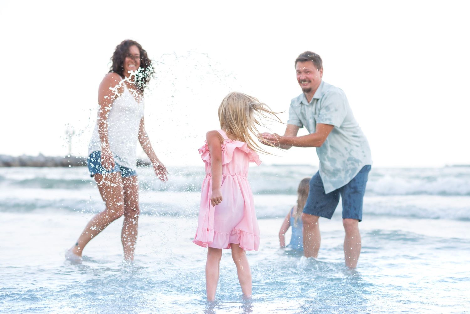 Family playing joyfully in ocean waves on a sunny beach, parents and children splashing in shallow water.