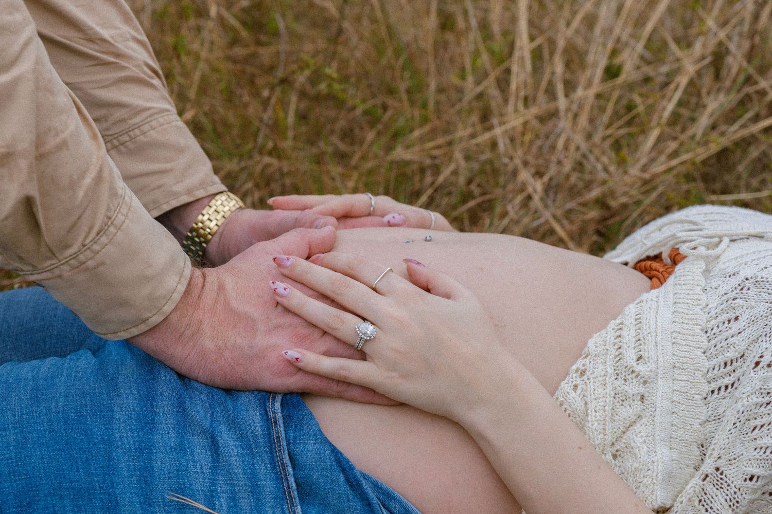 Couple tenderly placing hands on pregnant belly outdoors in a field, woman wearing engagement ring.