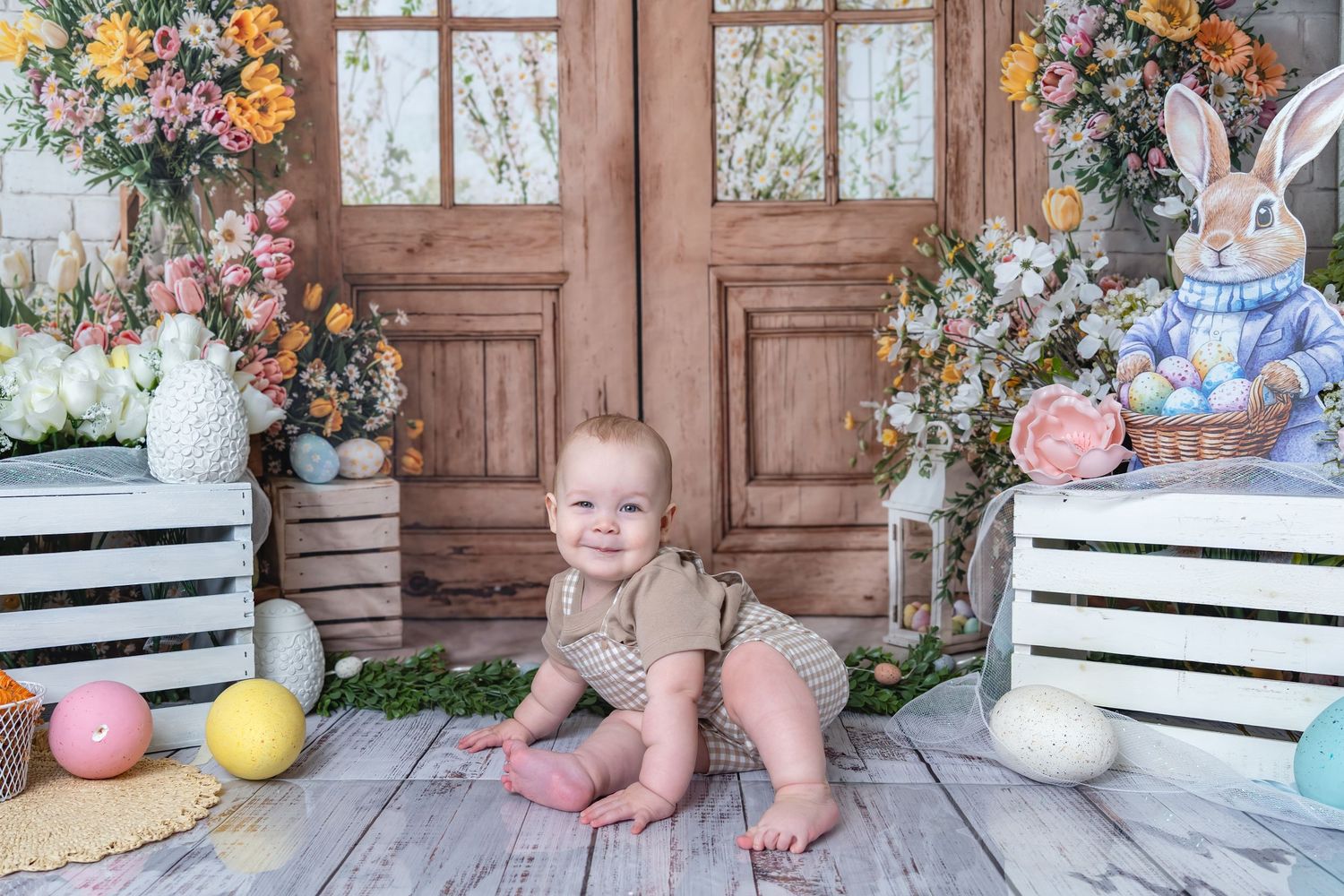 Baby in romper sits among Easter eggs, bunny decor, and spring flowers in a charming holiday photo setup.