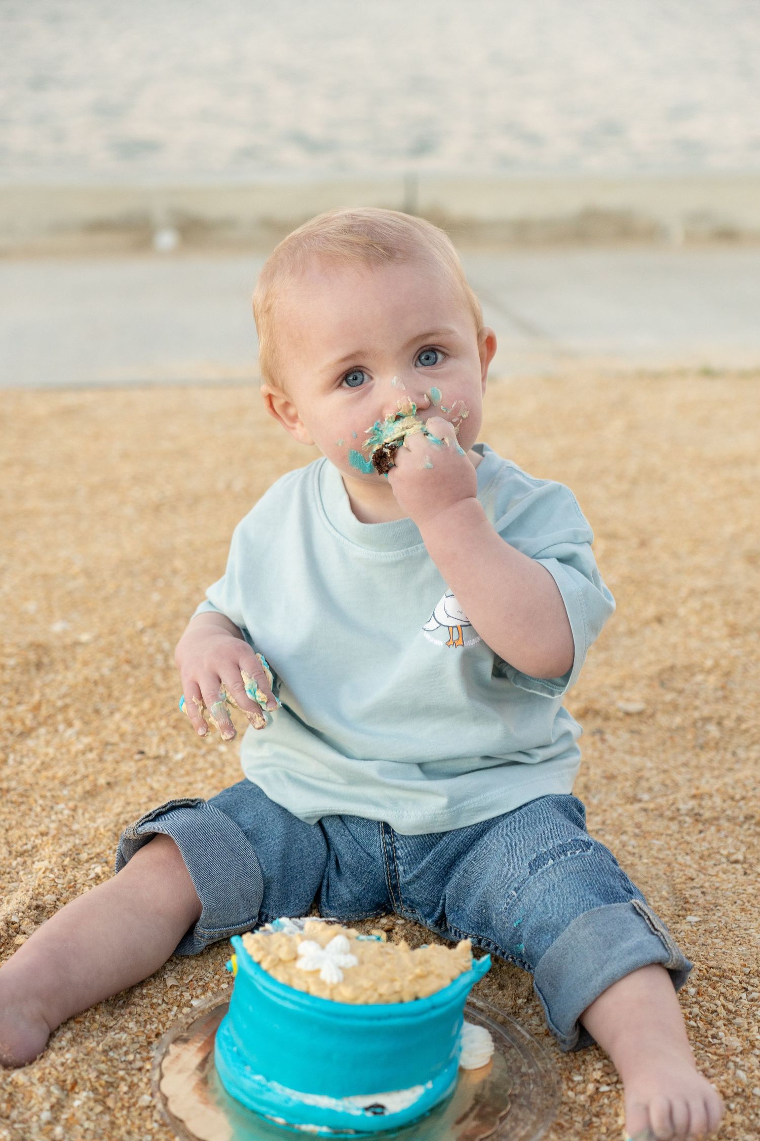 Baby boy sitting on beach sand eating smash cake for first birthday celebration outdoors.