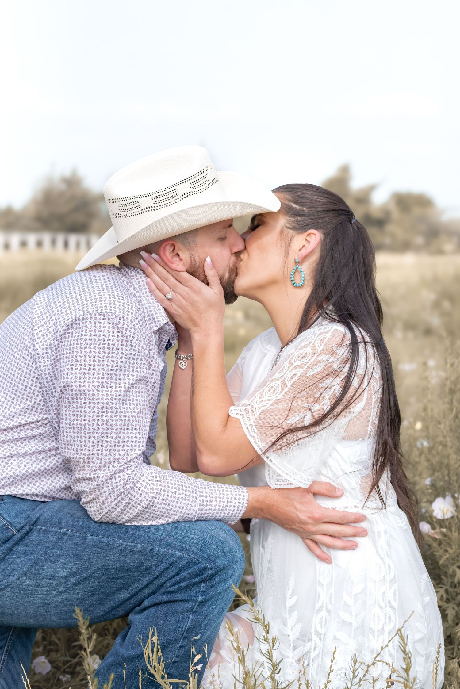 A couple sharing a romantic kiss in a field, man in cowboy hat and woman in white lace dress.