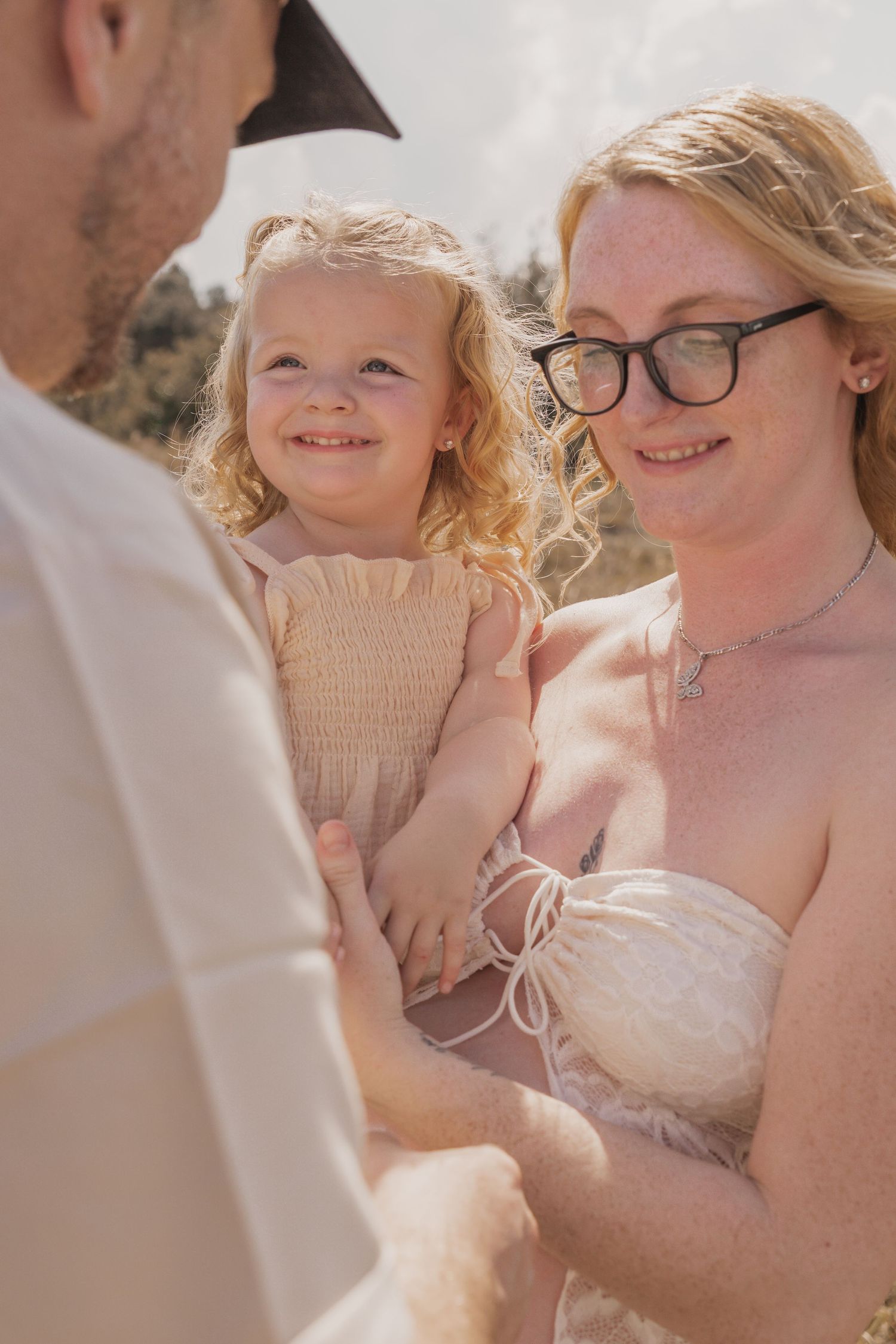 A smiling toddler girl with curly blonde hair held by her mother with glasses outdoors on a sunny day.