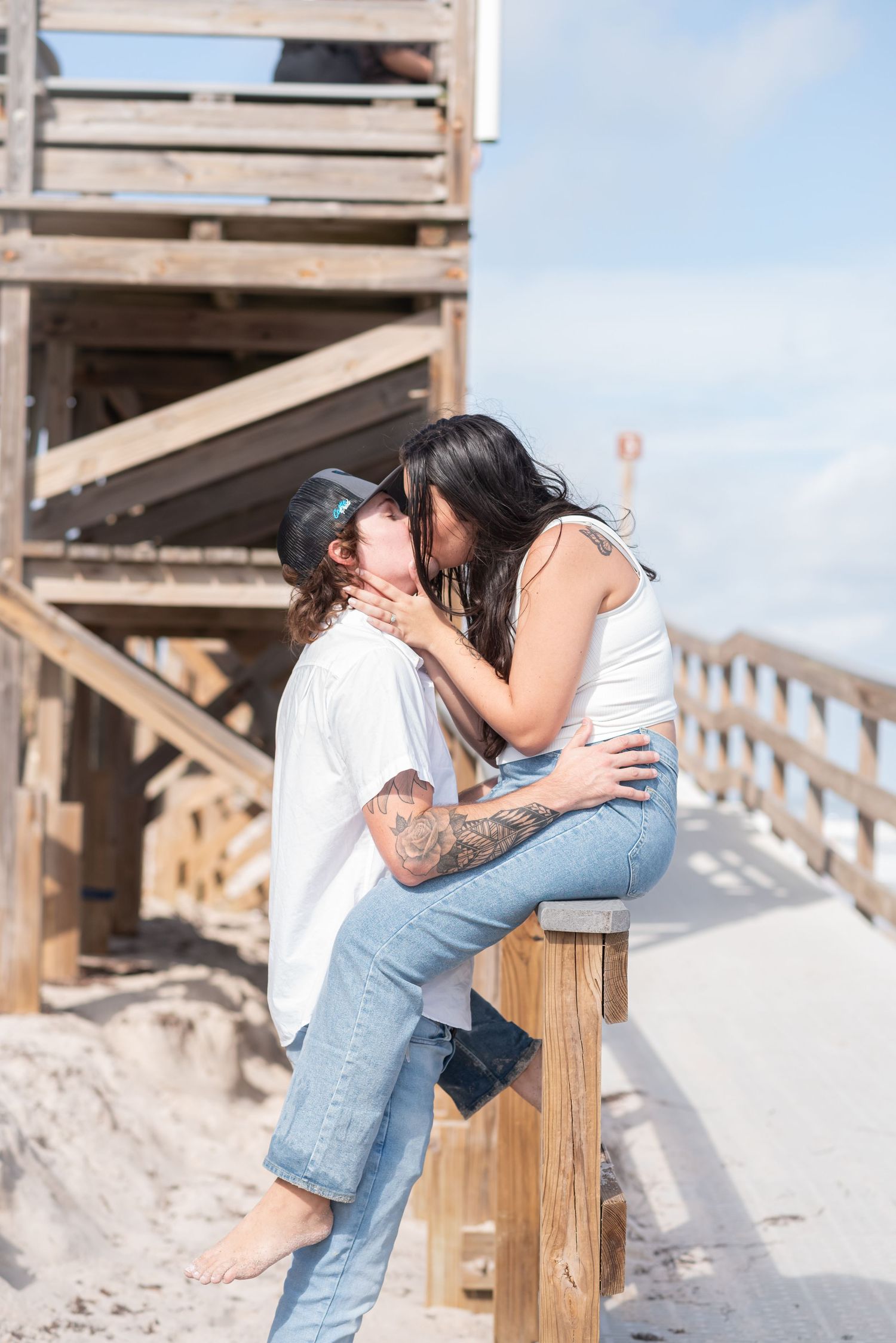 Couple sharing a romantic kiss near a beach lifeguard tower, wearing jeans and white tops on a sunny day.