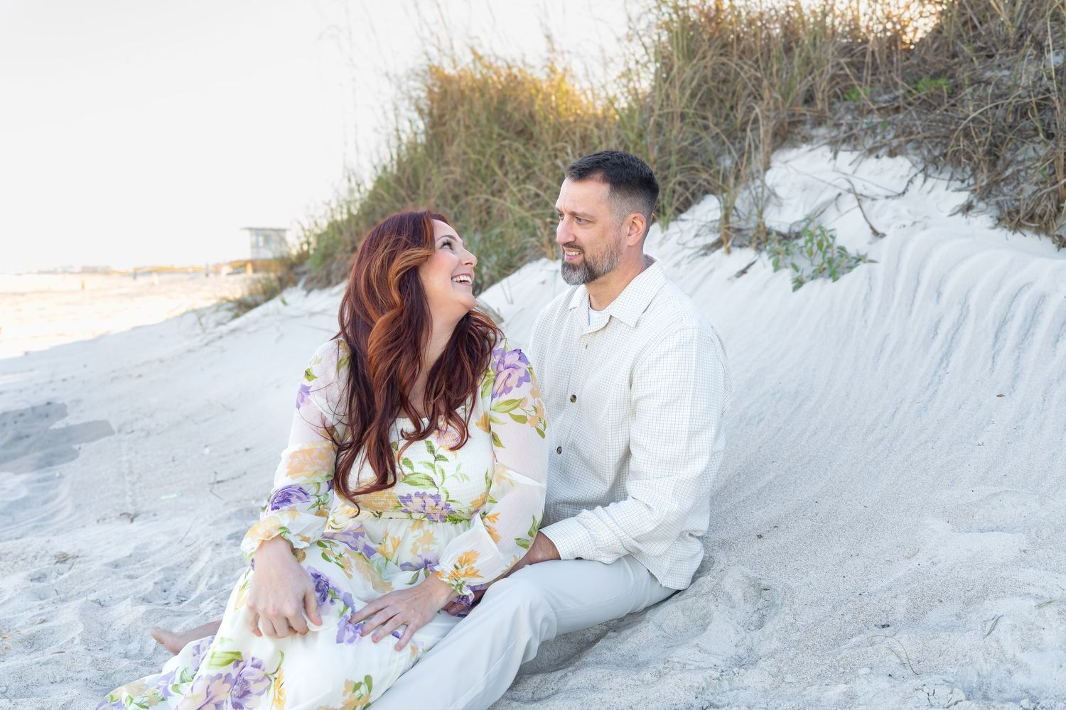 Couple sitting together on sandy beach dunes, woman in floral dress and man in white linen, sharing a joyful moment.