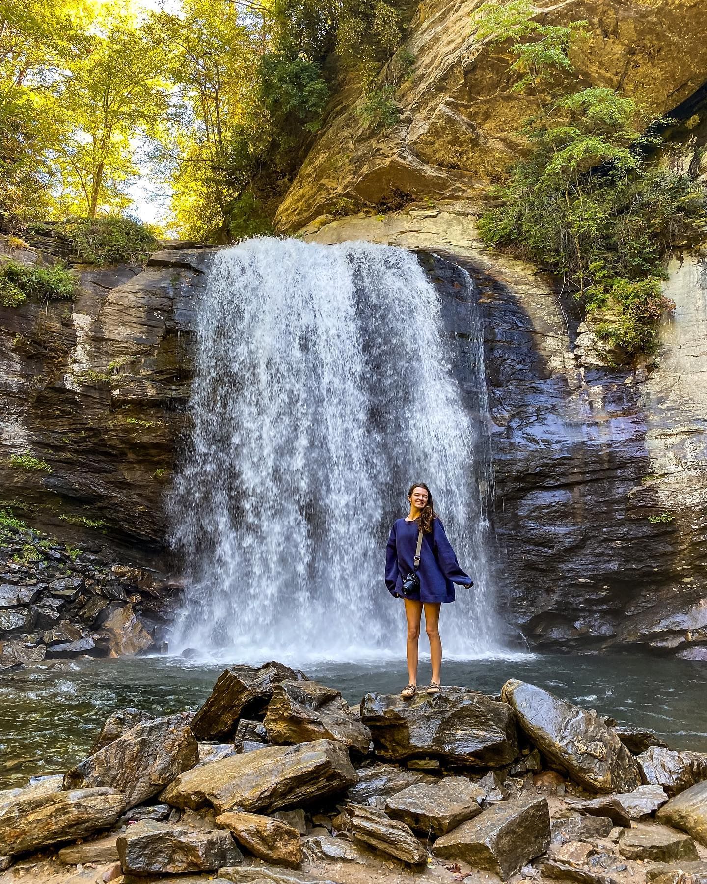 Woman in blue dress standing on rocks before tall cascading waterfall in forest.