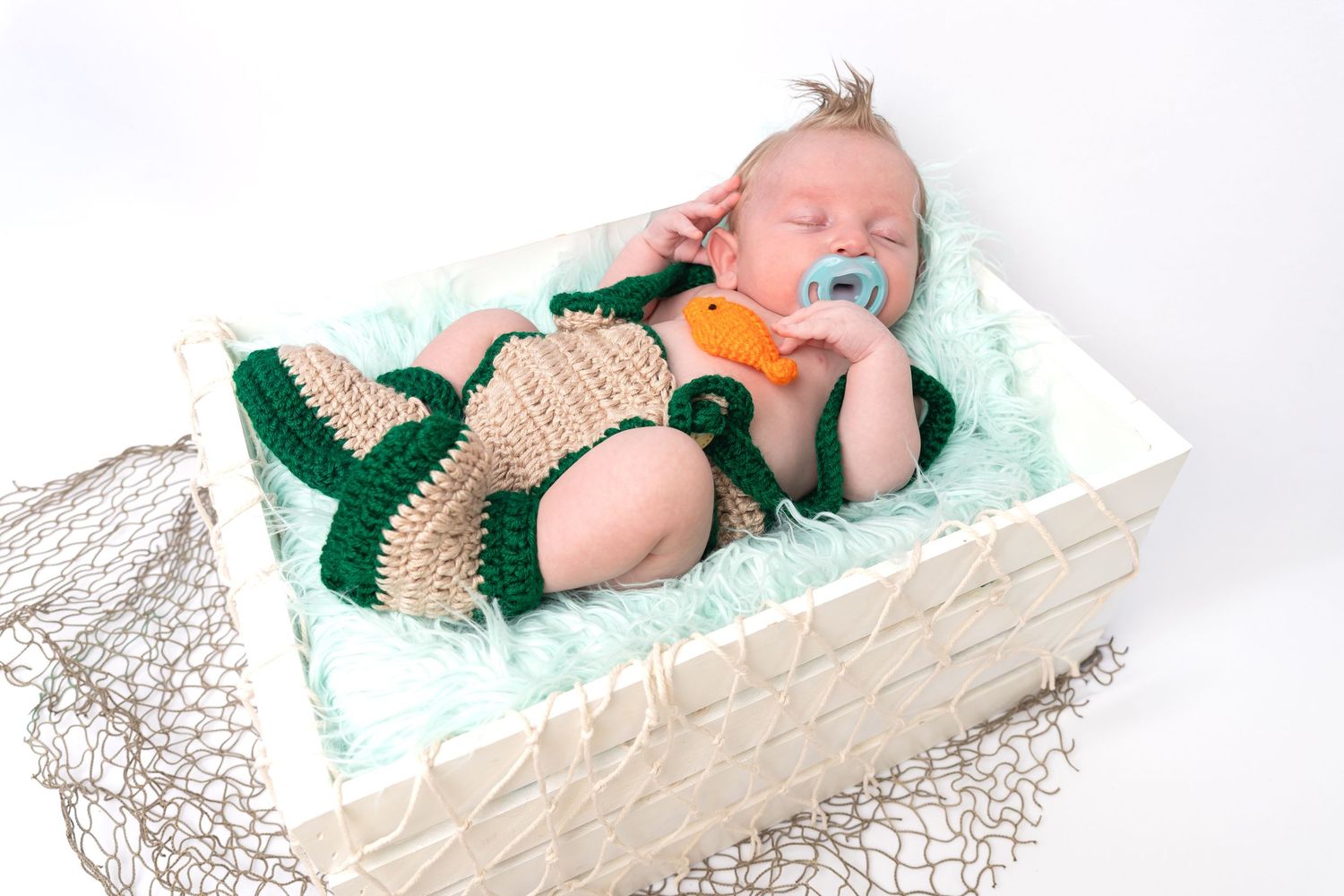 Sleeping newborn baby in crocheted fisherman outfit with orange pacifier, resting in white basket with green shredded paper.