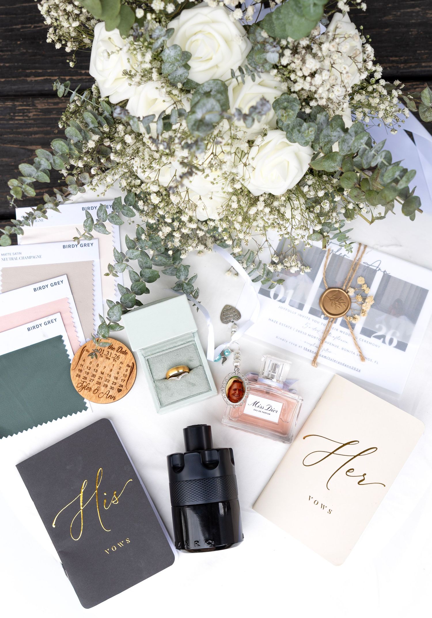Wedding flat lay with His and Hers vows books, ring box, Miss Dior perfume, cologne, and white rose bouquet.