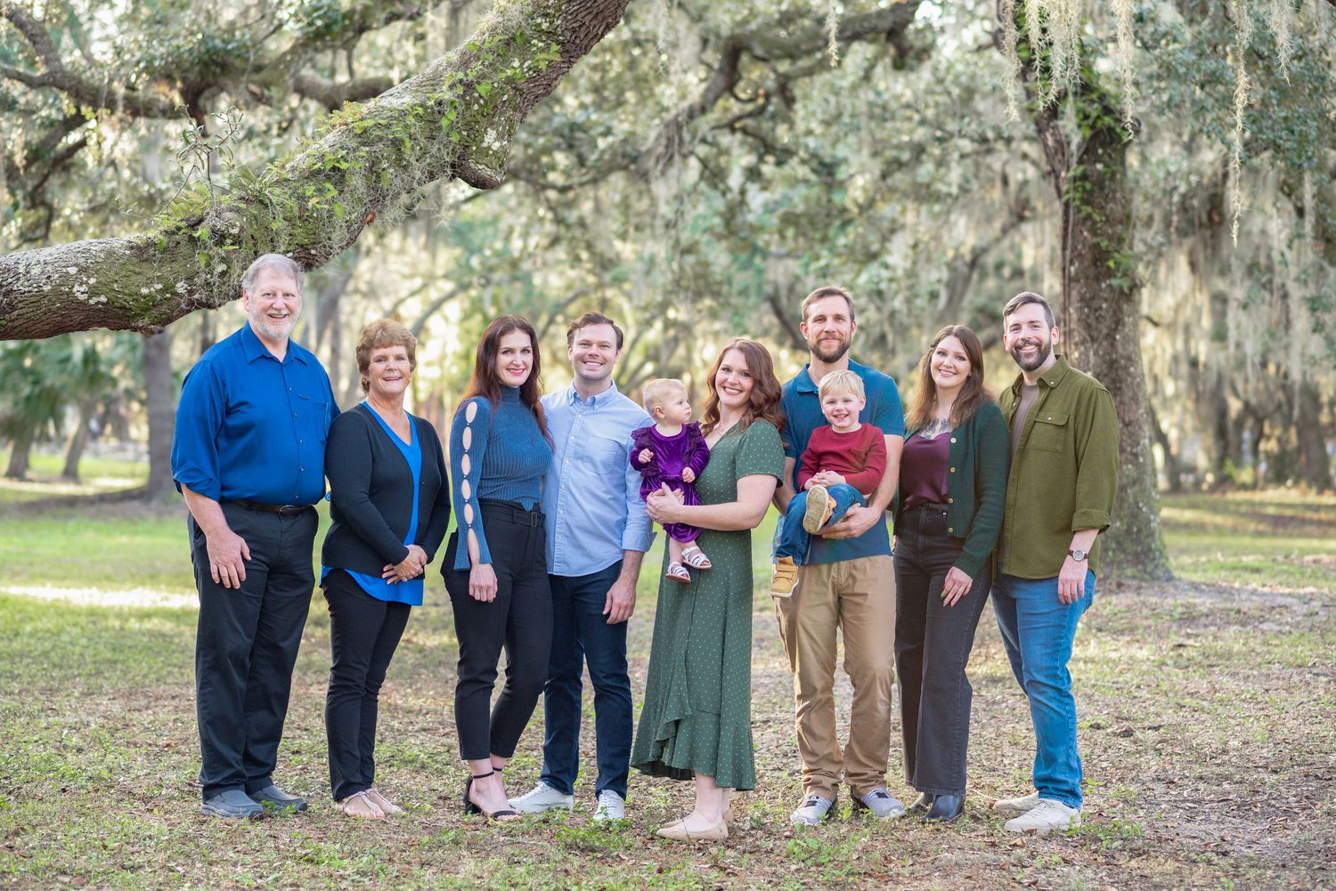 Large family group portrait outdoors under moss-covered oak trees, smiling together on a sunny day.