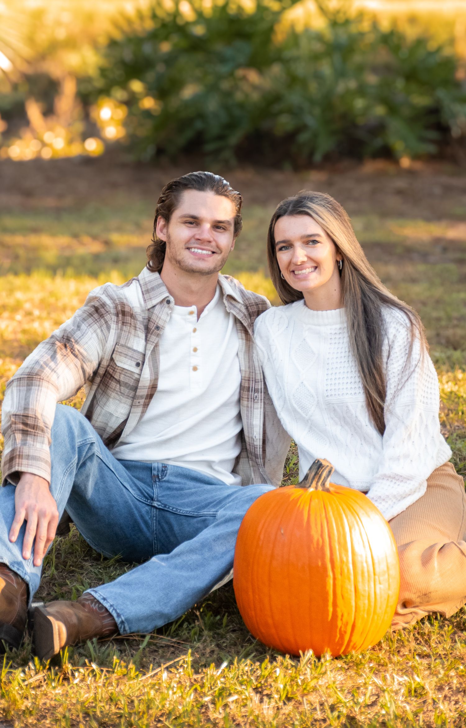 A smiling couple sits on grass with an orange pumpkin, dressed in fall attire, with greenery in the background.