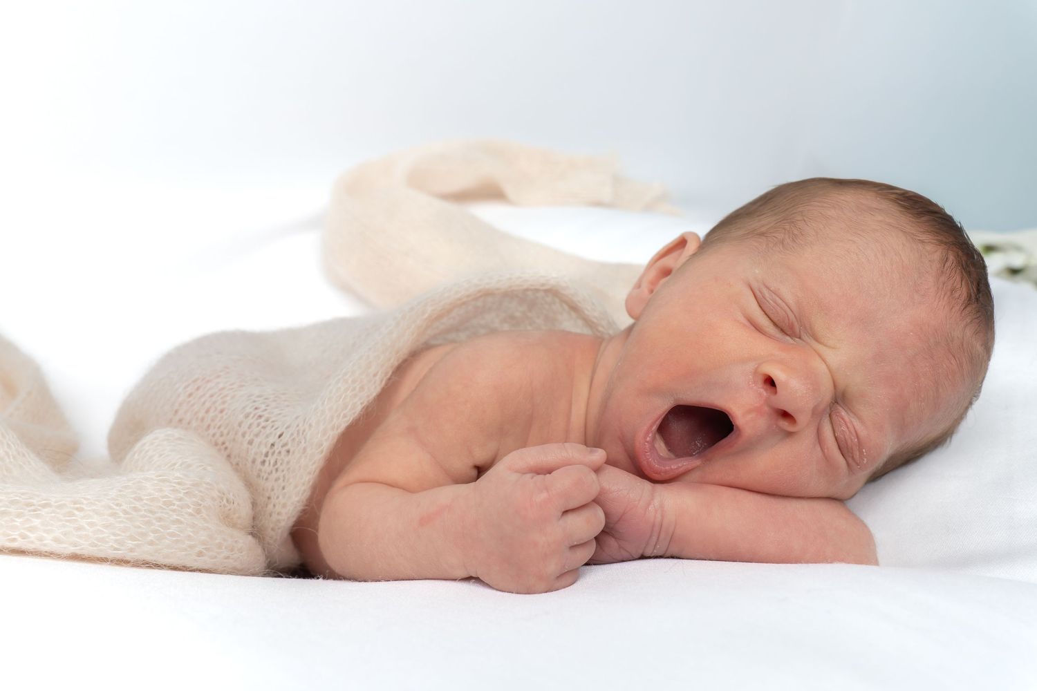 Adorable newborn baby yawning while lying on white blanket in soft natural light.