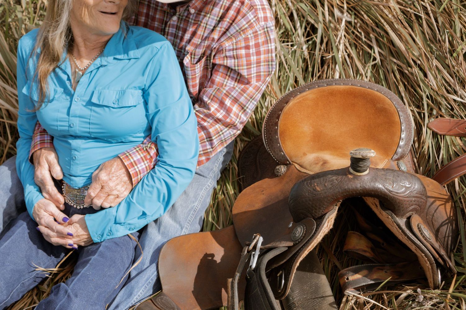 Two cowboys sitting in hay next to a leather western saddle, wearing plaid and blue shirts.