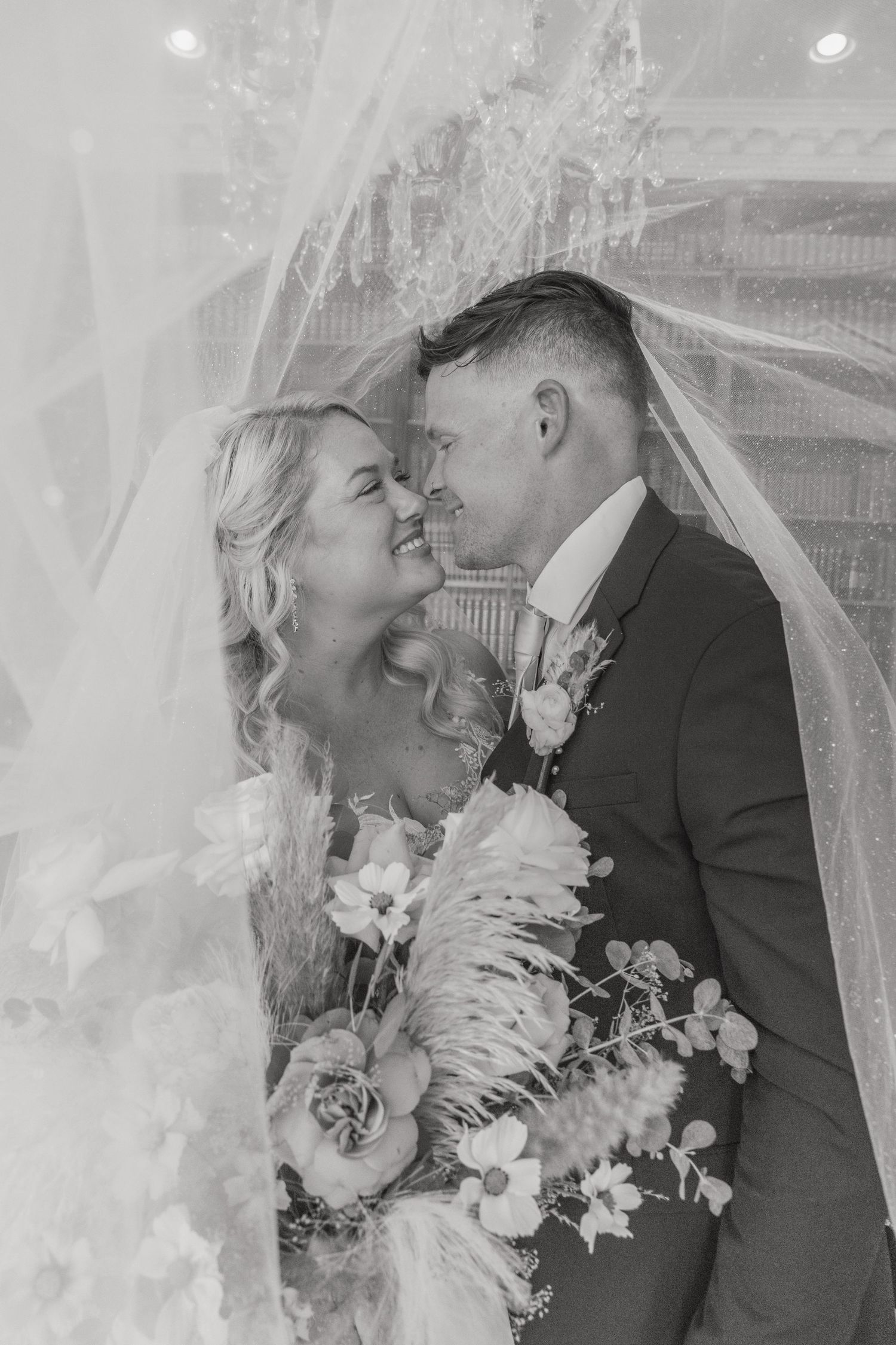 Black and white photo of smiling bride and groom embracing under a flowing veil on their wedding day.