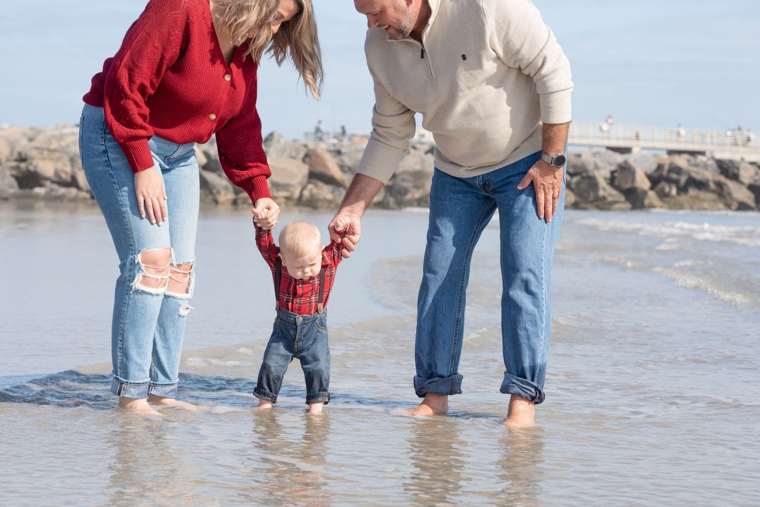 Family helping baby take first steps on a sunny beach, parents holding toddler's hands at water's edge.