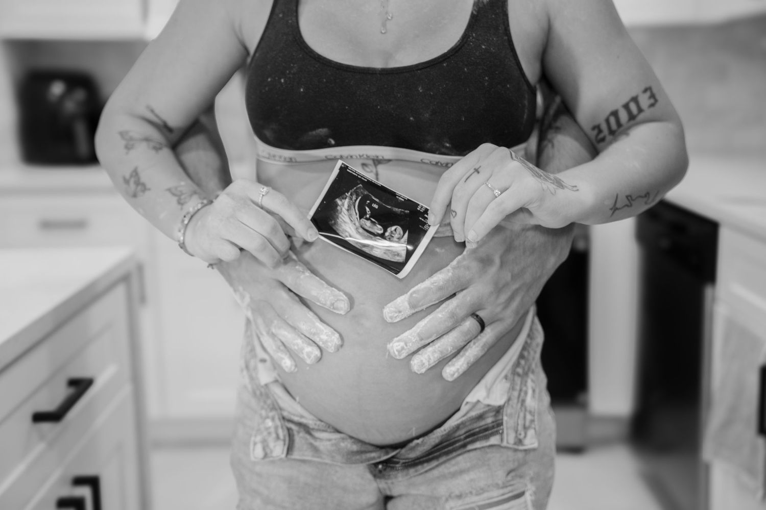 Pregnant woman holding ultrasound photo against baby bump, black and white maternity photography with tattoos visible.