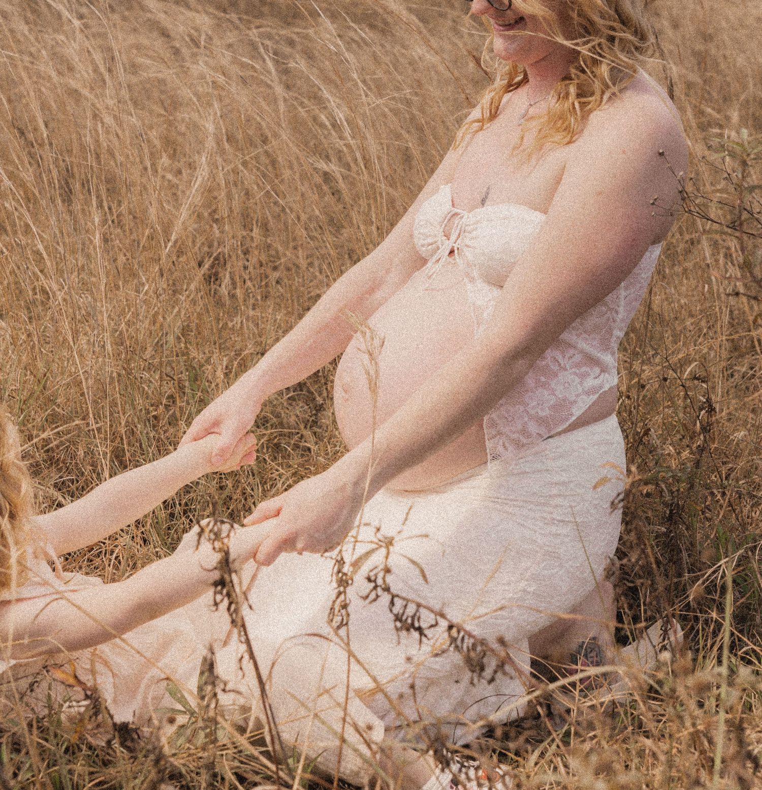 Pregnant woman in white lace dress sitting in golden grass field, holding child's hand during maternity photoshoot.