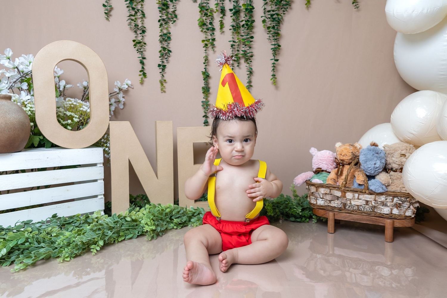 Baby's first birthday cake smash photo with ONE letters, party hat, balloons, and stuffed animals on beige backdrop.