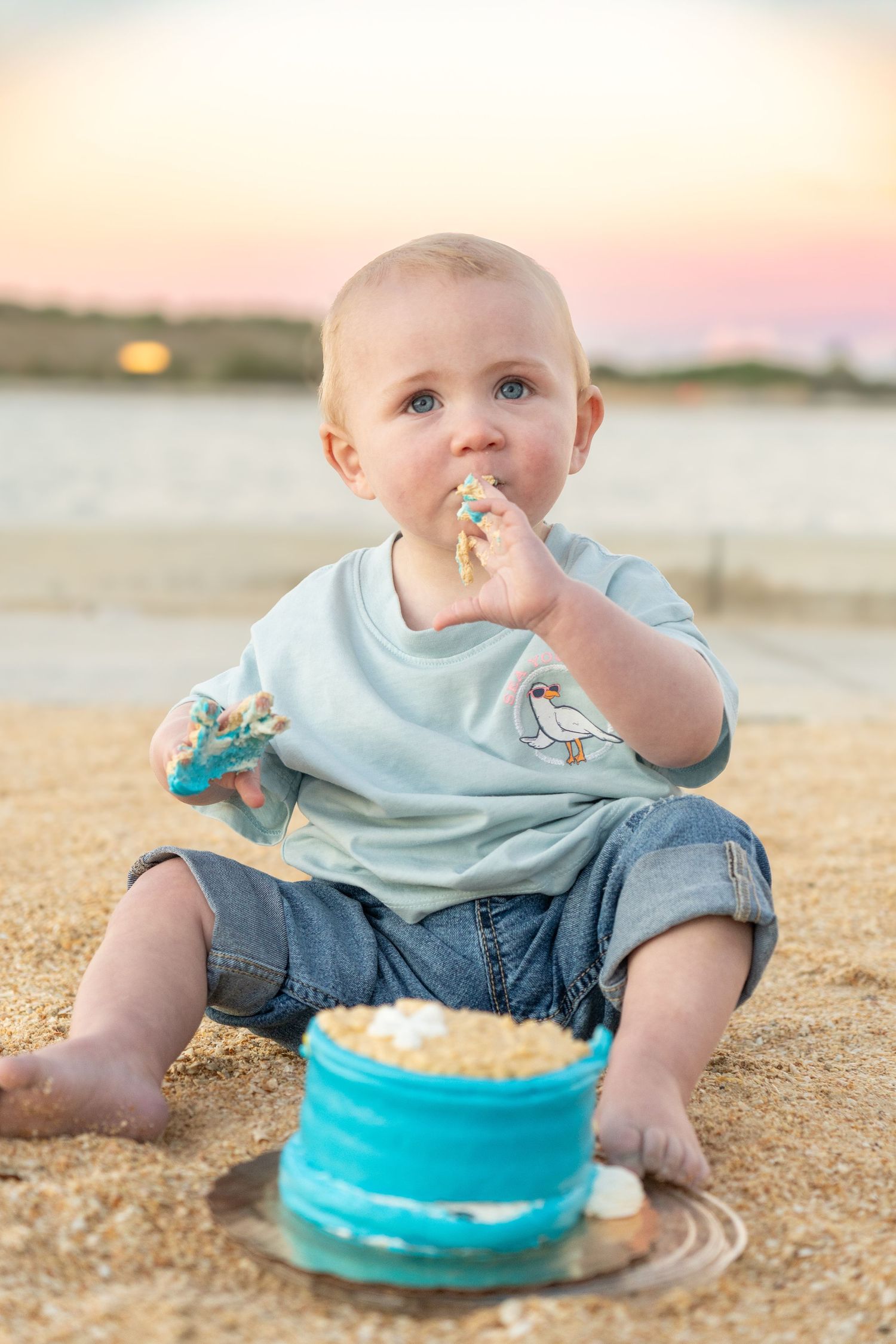 Baby boy eating smash cake on sandy beach at sunset, wearing light blue shirt and denim shorts.