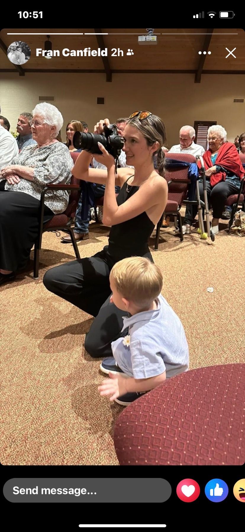 Female photographer crouching to capture photos at an indoor event with elderly audience members seated nearby.