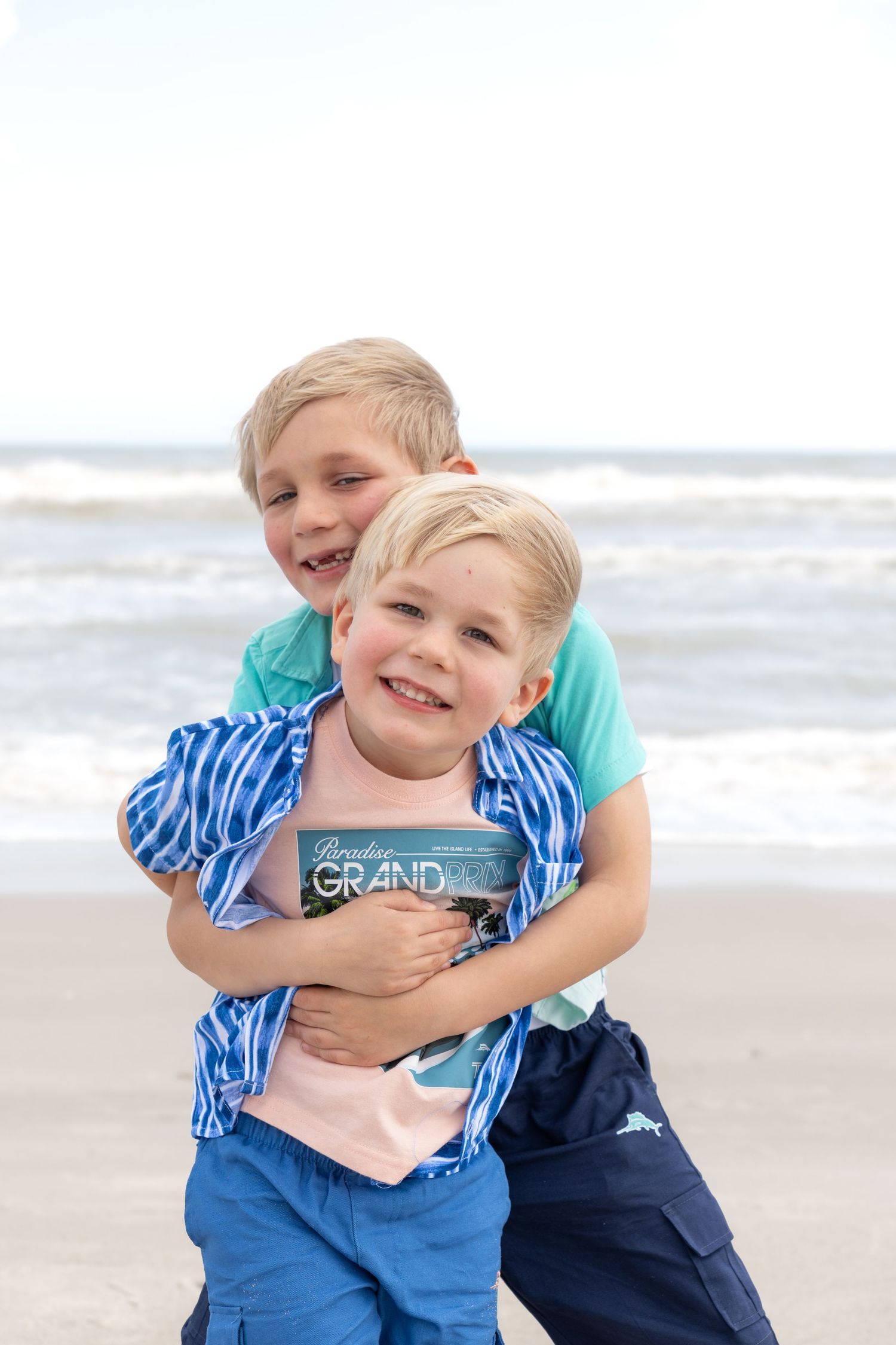 Two blonde boys hugging and smiling on a beach with ocean waves in the background.