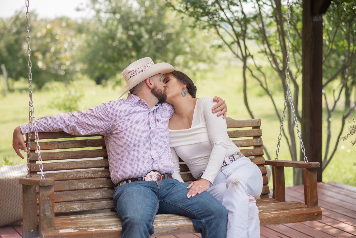 Couple sharing a tender kiss on a wooden porch swing surrounded by lush green trees.