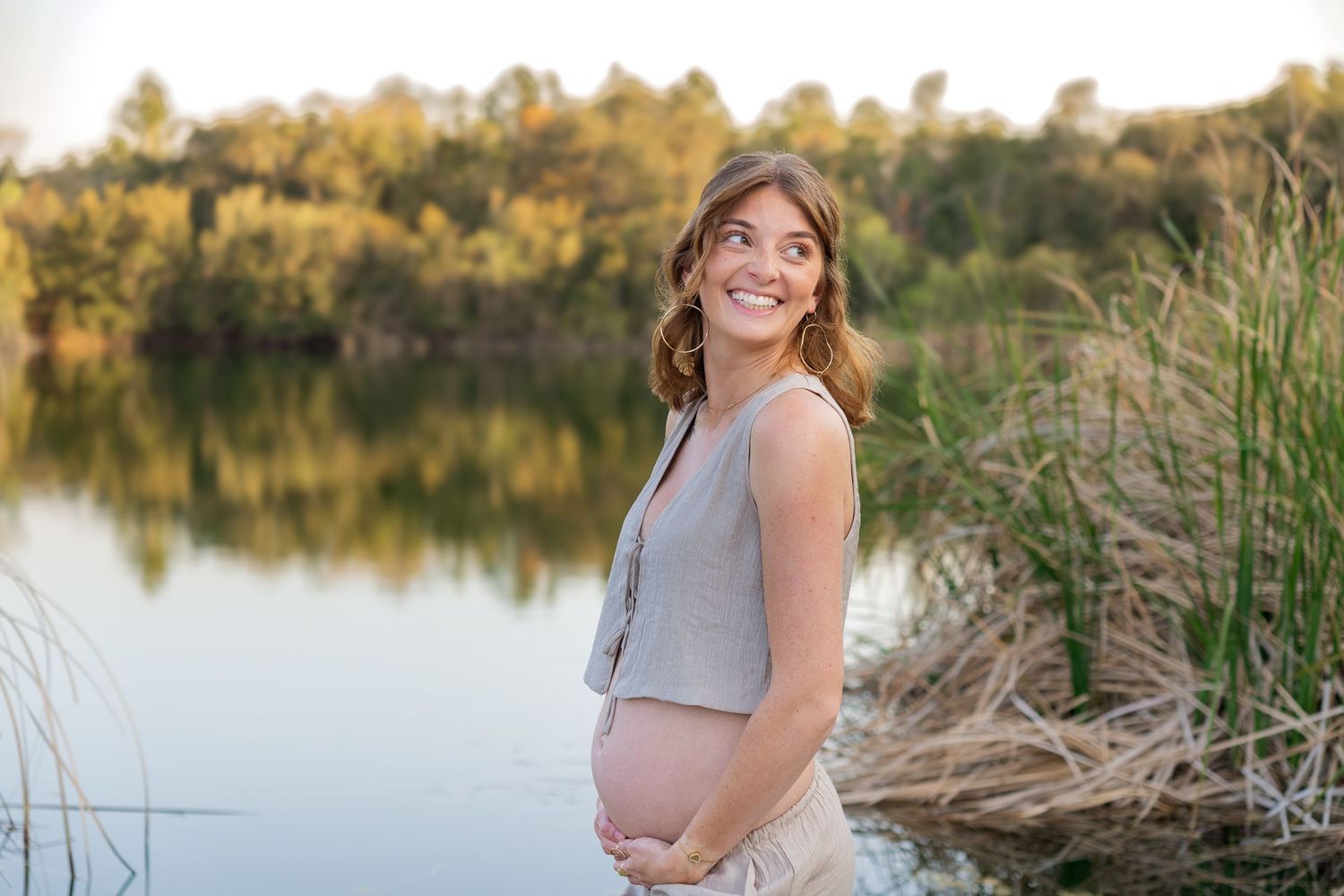 Smiling pregnant woman cradling her baby bump by a serene lake surrounded by lush greenery during golden hour.