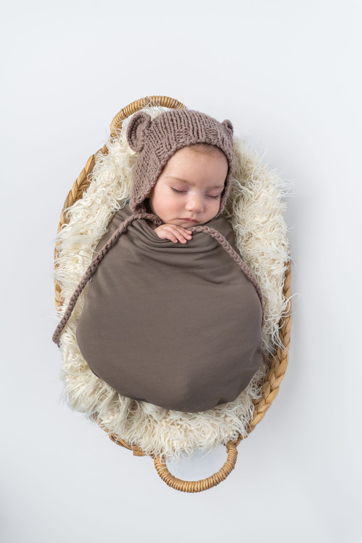 Sleeping newborn baby wrapped in brown swaddle wearing knit bear bonnet in wicker basket with fluffy lining.