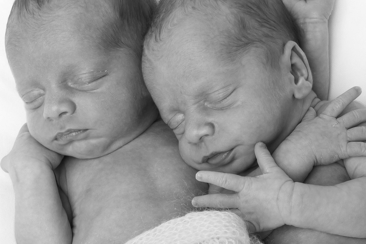 Black and white photo of two sleeping newborn twins snuggled closely together, holding hands.