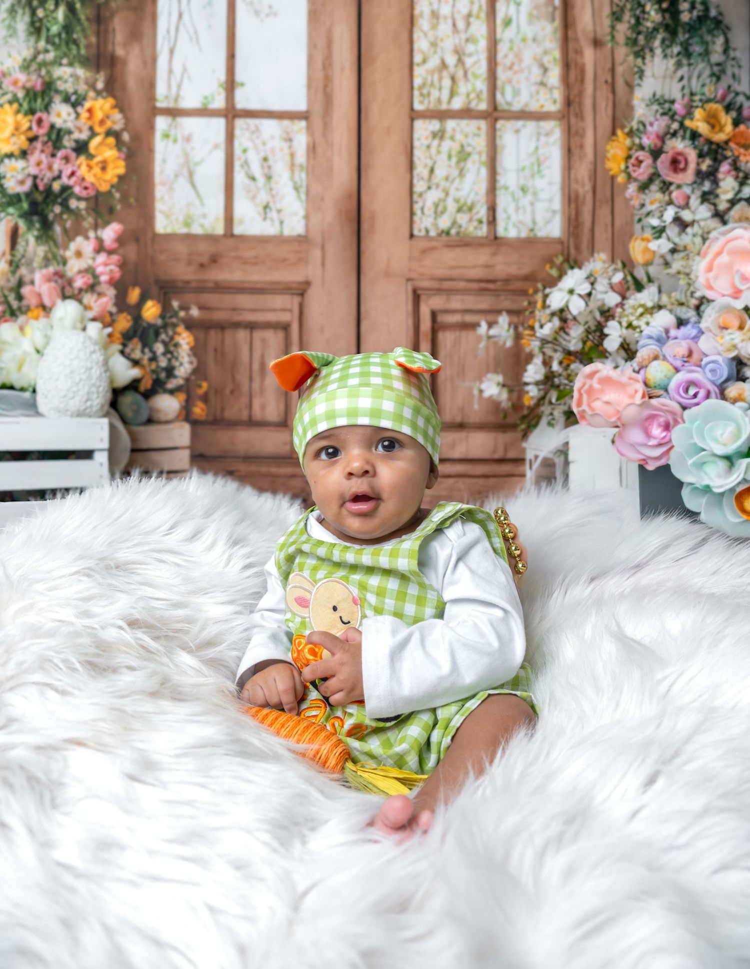 Baby in green gingham romper and hat sits on white fur rug surrounded by colorful spring flowers and wooden doors.