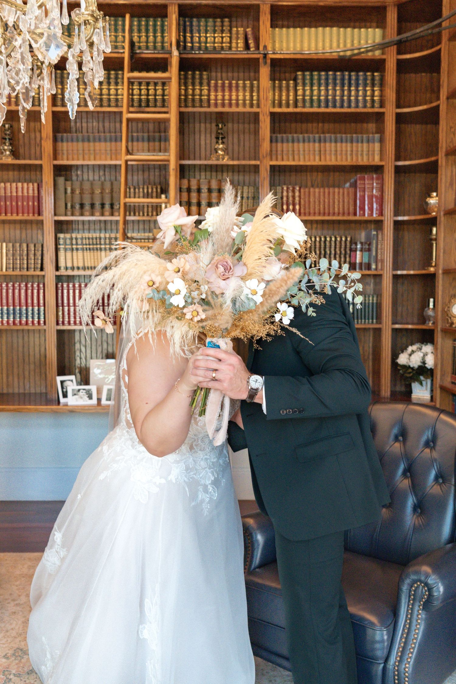 Bride and groom kissing behind a boho bouquet of pampas grass and roses in an elegant library setting.