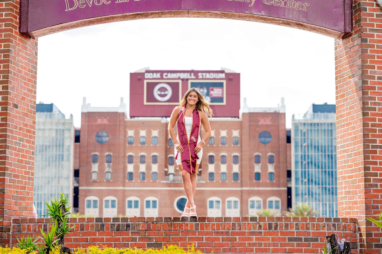 Young woman in maroon graduation dress posing under brick arch at Doak Campbell Stadium.