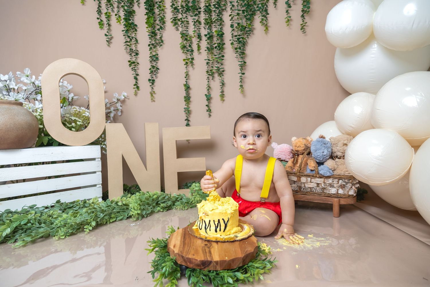 Whimsical first birthday setup with balloon arch, wooden decorations and greenery backdrop.