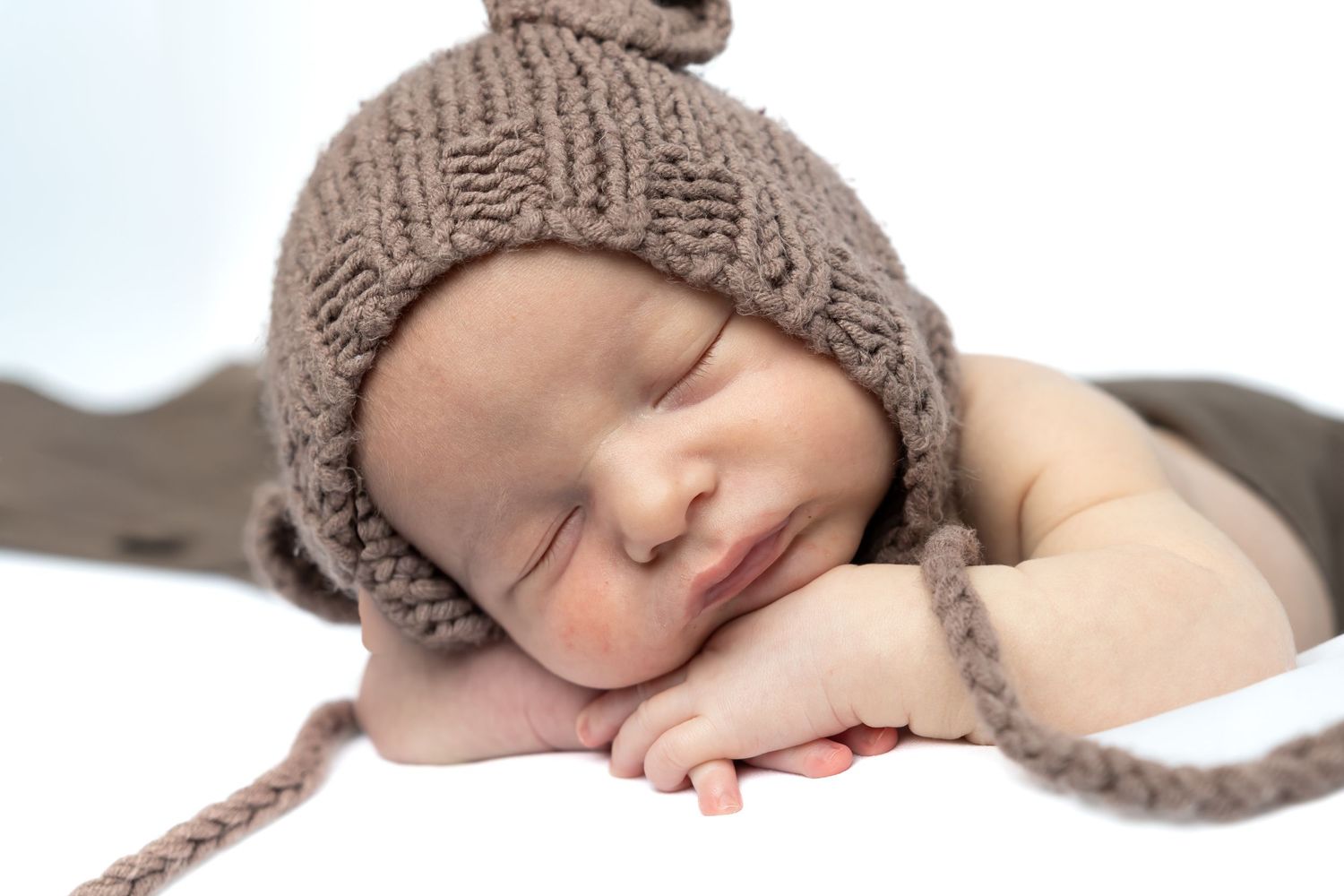 Sleeping newborn baby wearing a brown knitted hat with earflaps resting on tiny hands.