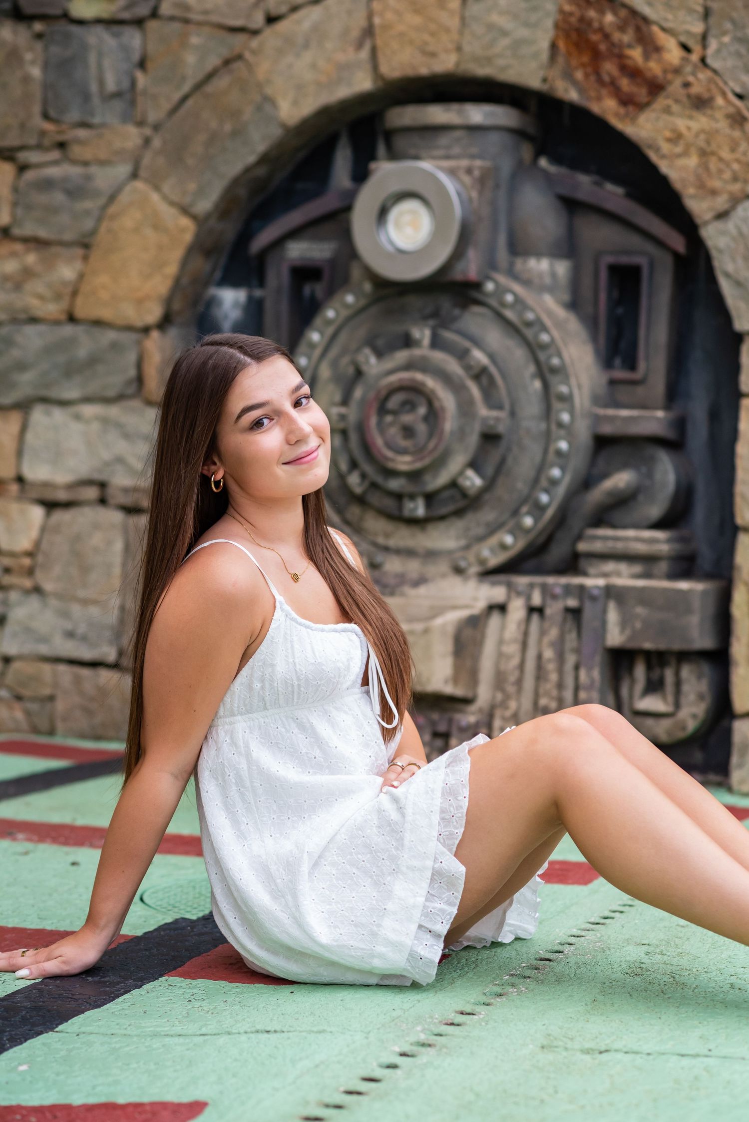 Young woman in white sundress sitting on colorful floor in front of ornate stone vault door backdrop.