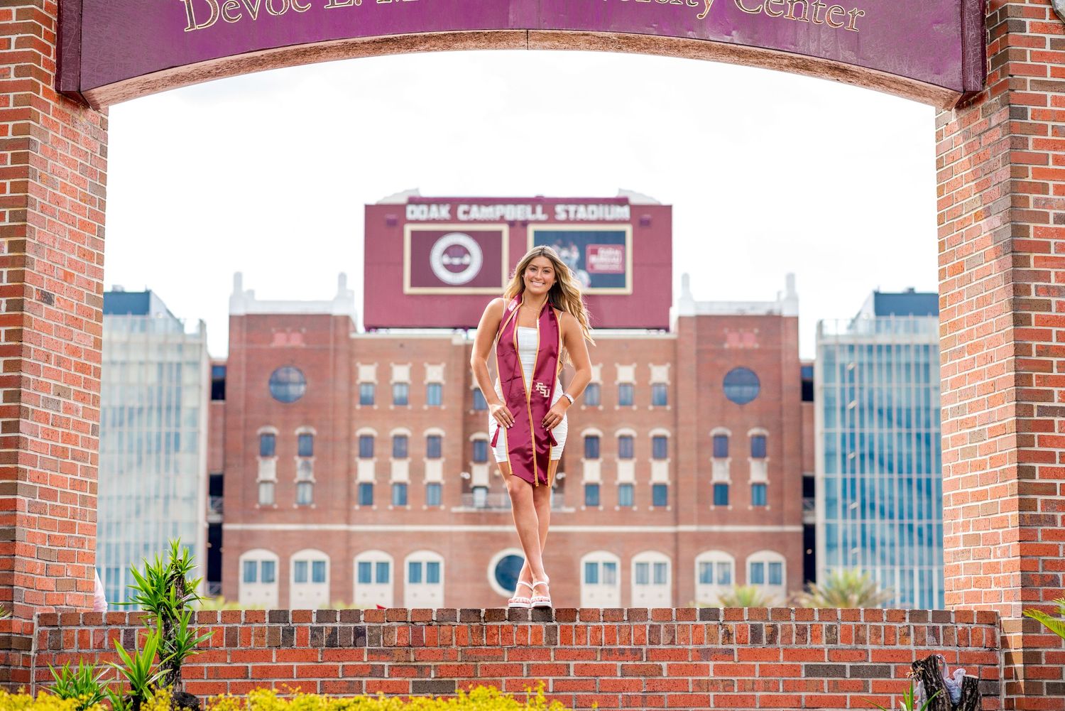 Graduate poses under brick arch at Doak Campbell Stadium in maroon and gold regalia.