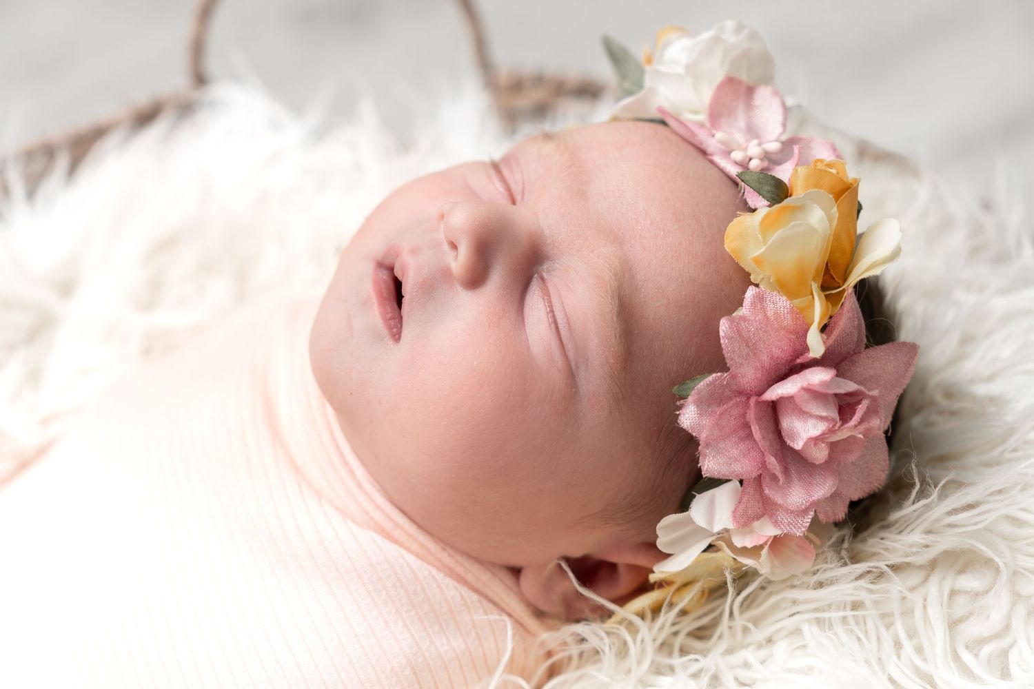 Sleeping newborn baby wearing a delicate floral headband with pink and gold flowers, wrapped in soft pink fabric.