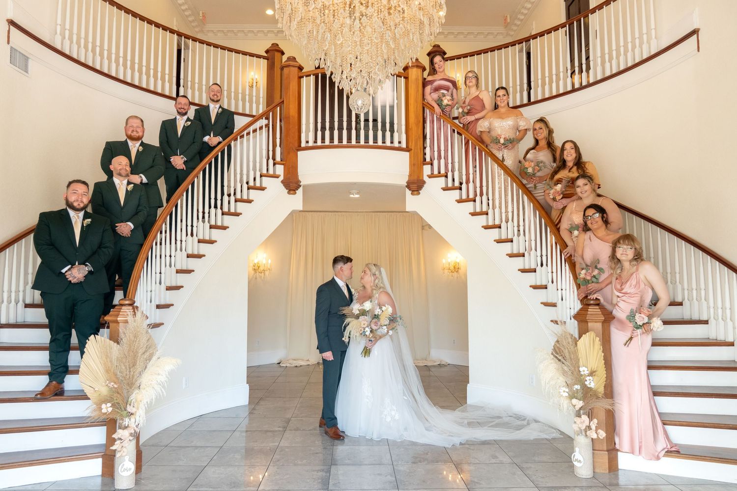 Wedding party poses on grand double staircase with chandelier, bride and groom centered, groomsmen and bridesmaids on steps.